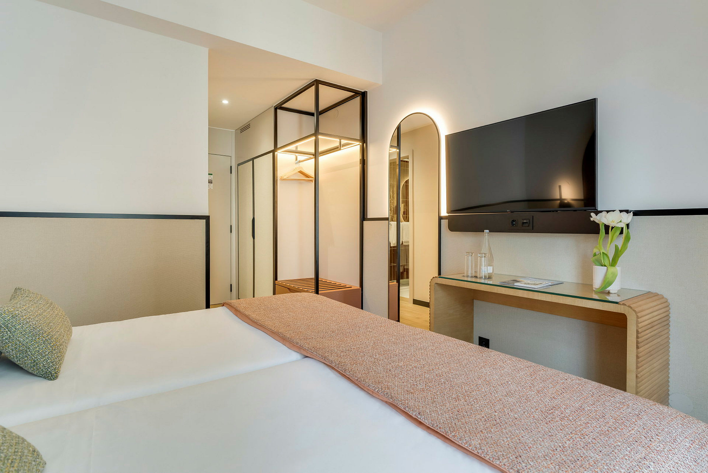 Ett hotellrum med en dubbelsäng, en spegel, en TV och en skrivbord med en vas med vita blommor. (AI-genererad text)