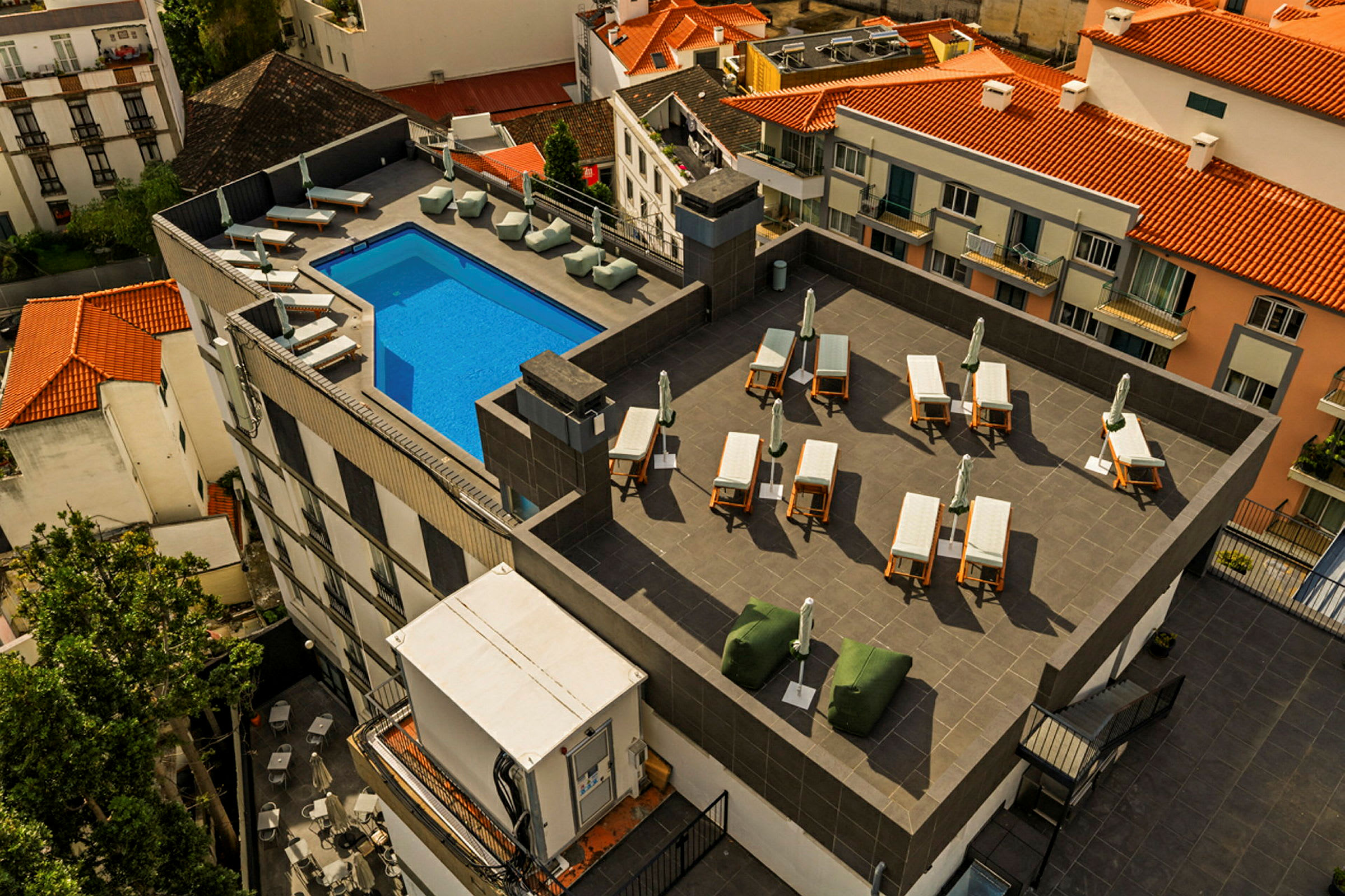 Bild av en takterrass med en blå pool, solstolar och parasoller, omgiven av byggnader med tegeltak. (AI-genererad text)