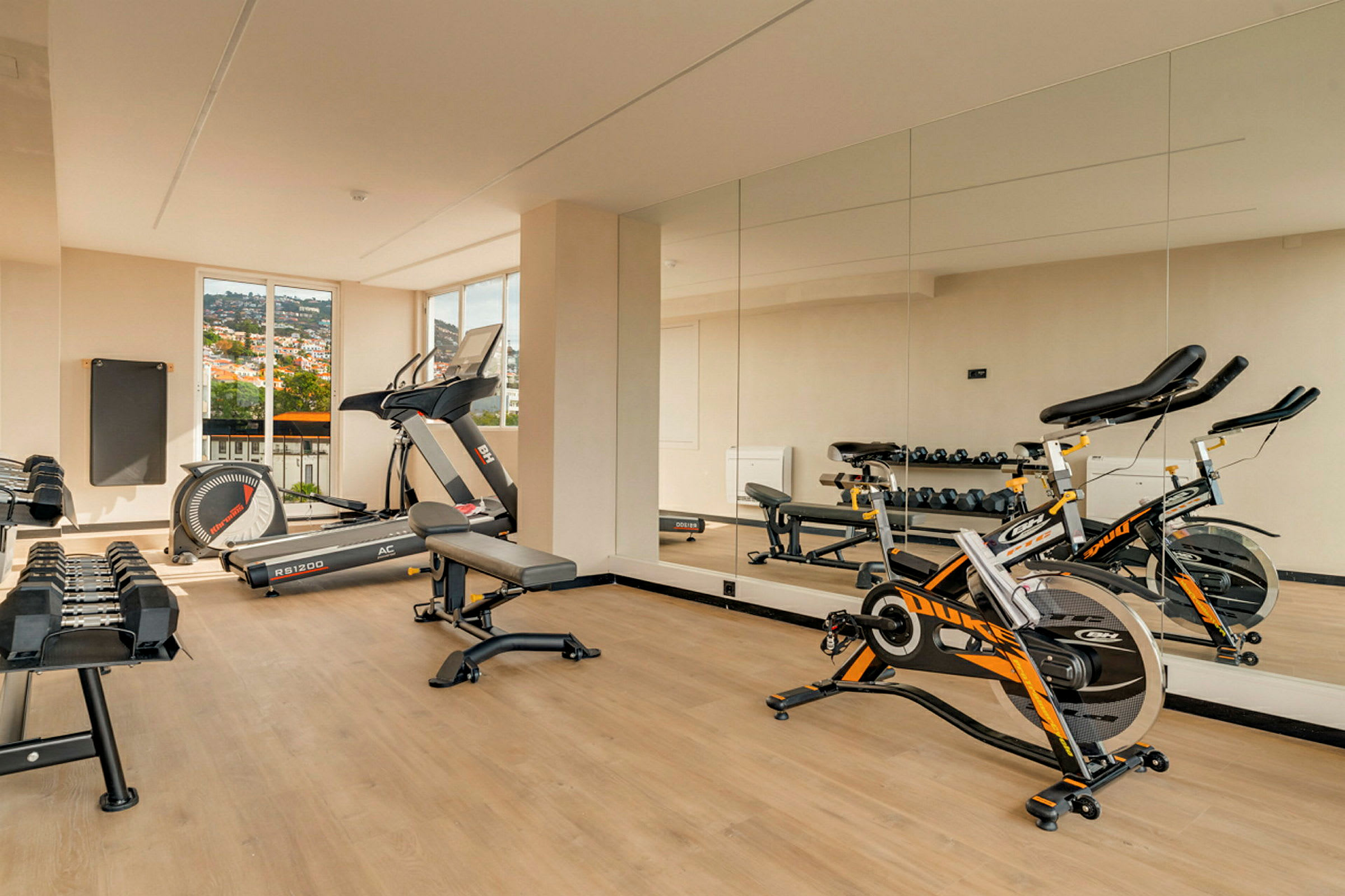 Ett gym med träningsutrustning som löpband, cykel och vikter, med spegelklädda väggar och fönster som visar utsikten utanför. (AI-genererad text)