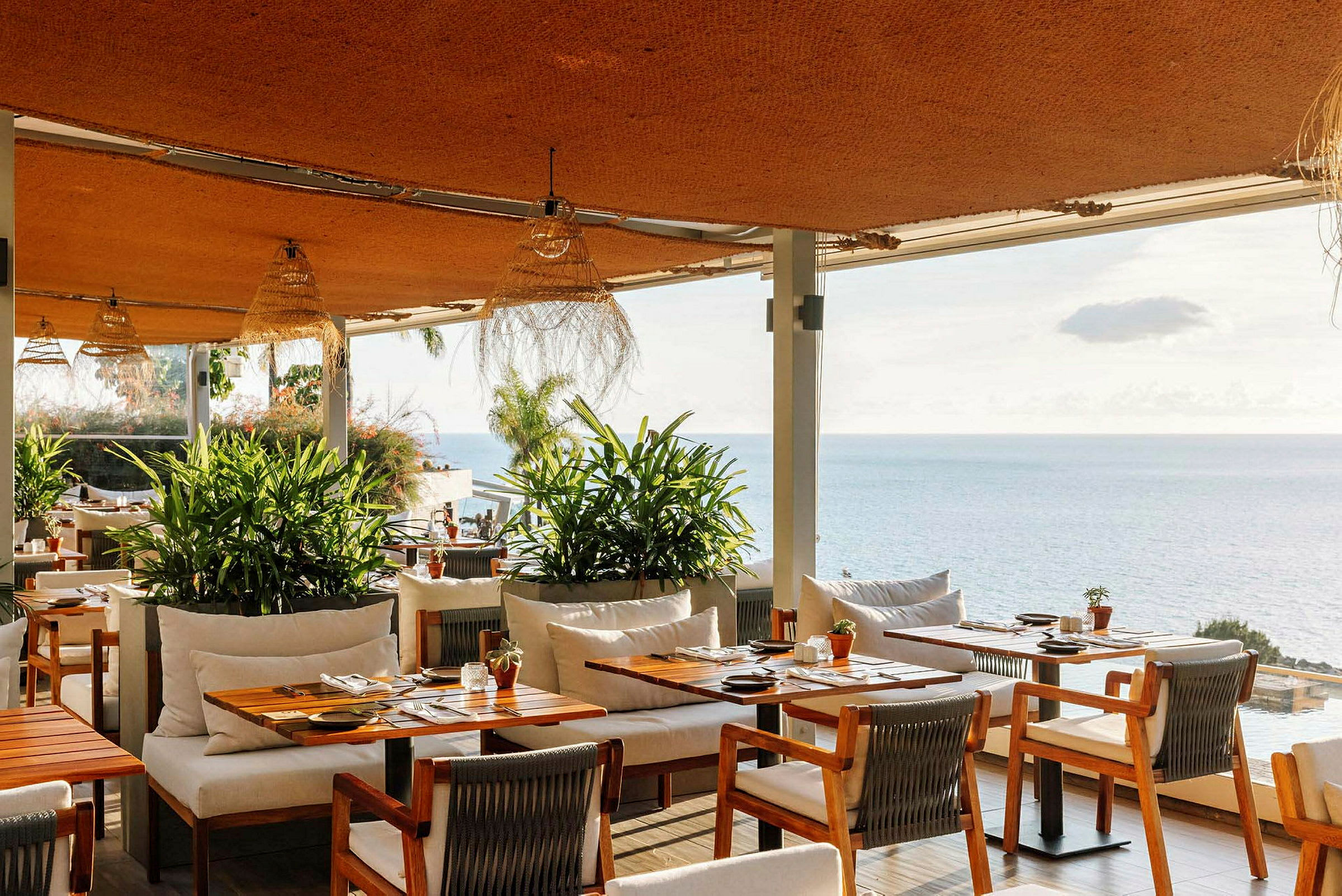 Utsikt över en restaurang med träbord och stolar, gröna växter och havet i bakgrunden. (AI-genererad text)