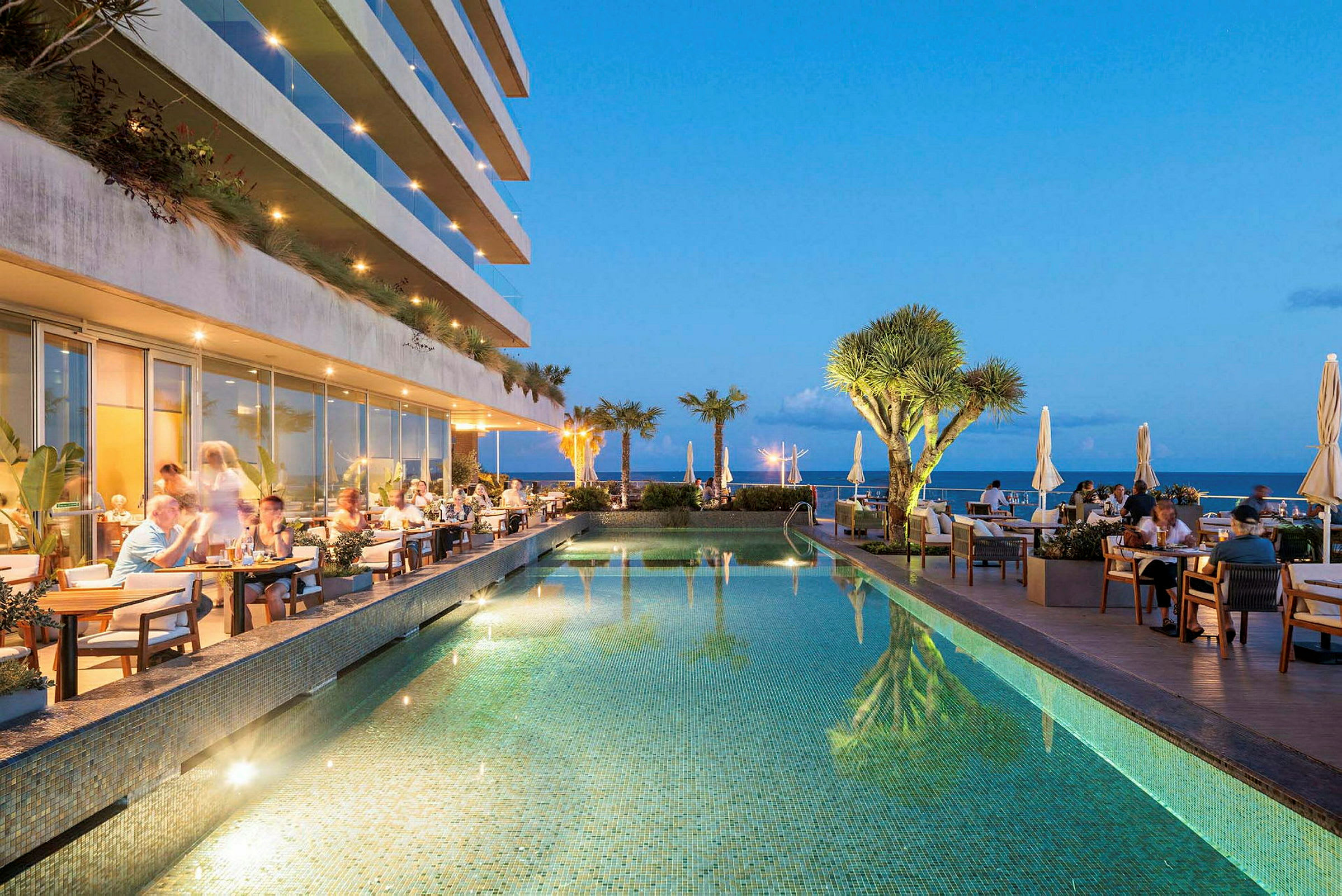 En restaurang med sittplatser vid sidan av en pool, med utsikt över havet och omgivande växter. (AI-genererad text)