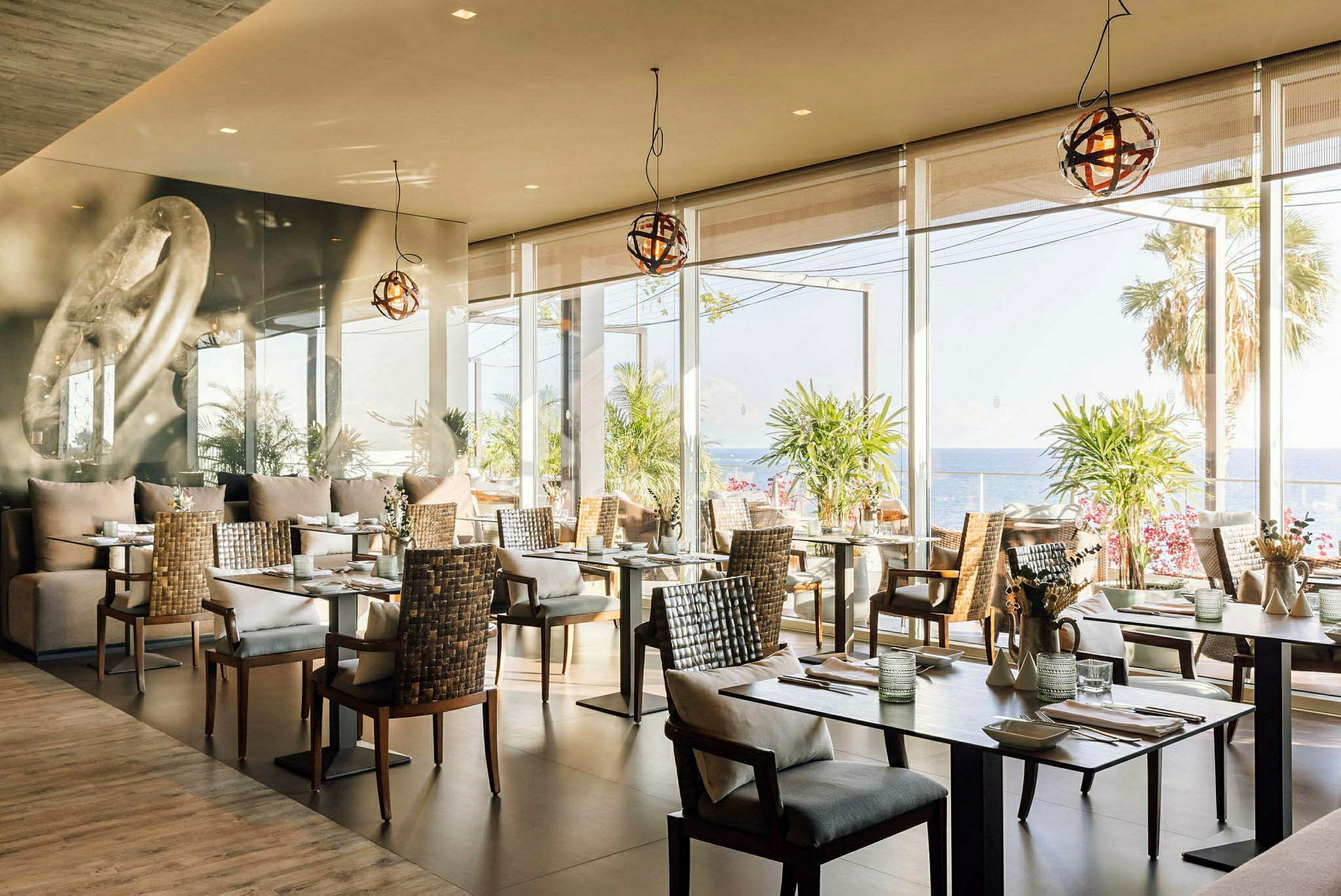 En restaurang med moderna bord och stolar, stora fönster som visar utsikt över havet och palmträd. (AI-genererad text)