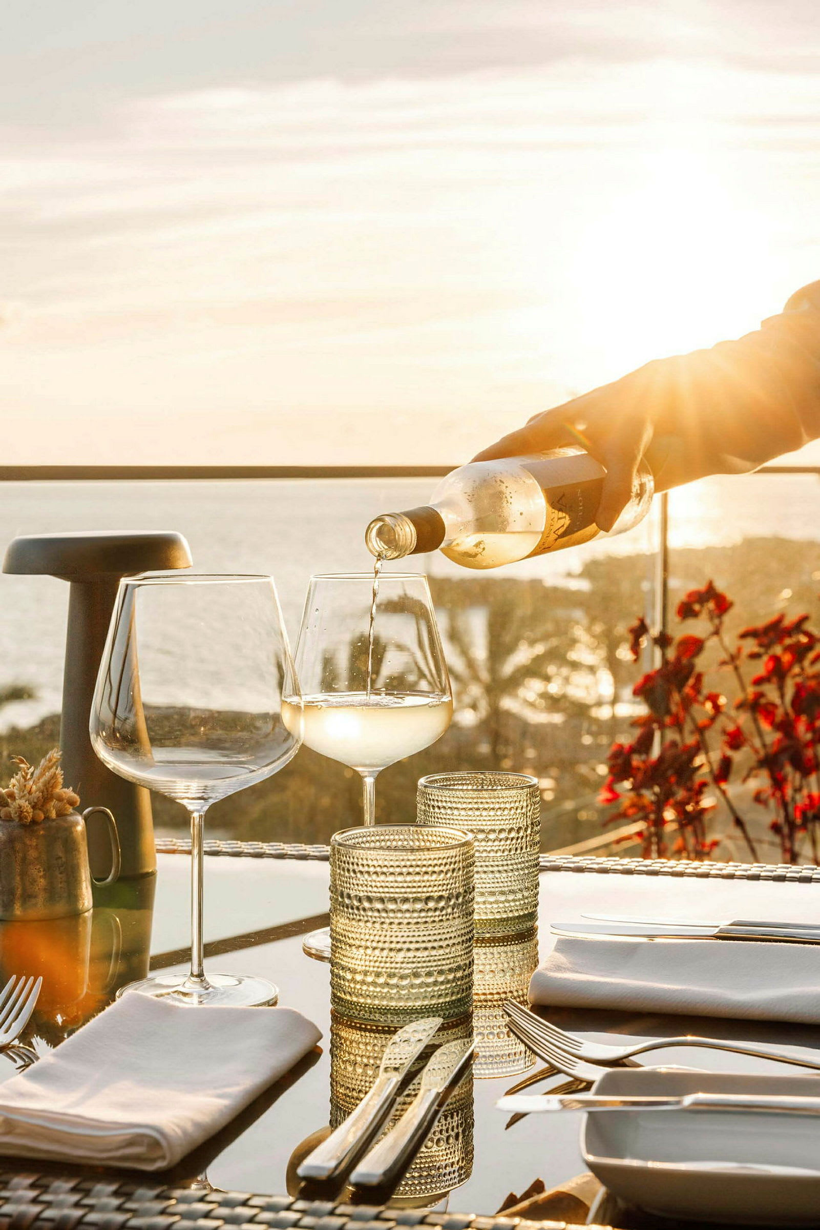 Vit dryck hälls upp i ett vinglas på ett bord med redskap och glas, med solnedgång och hav i bakgrunden. (AI-genererad text)