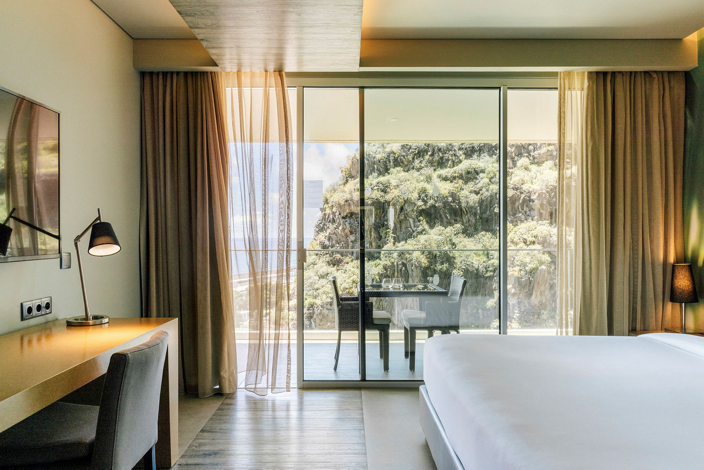 Motiv föreställer ett hotellrum med skrivbord, stol och säng, med utsikt mot en balkong och berglandskap. (AI-genererad text)