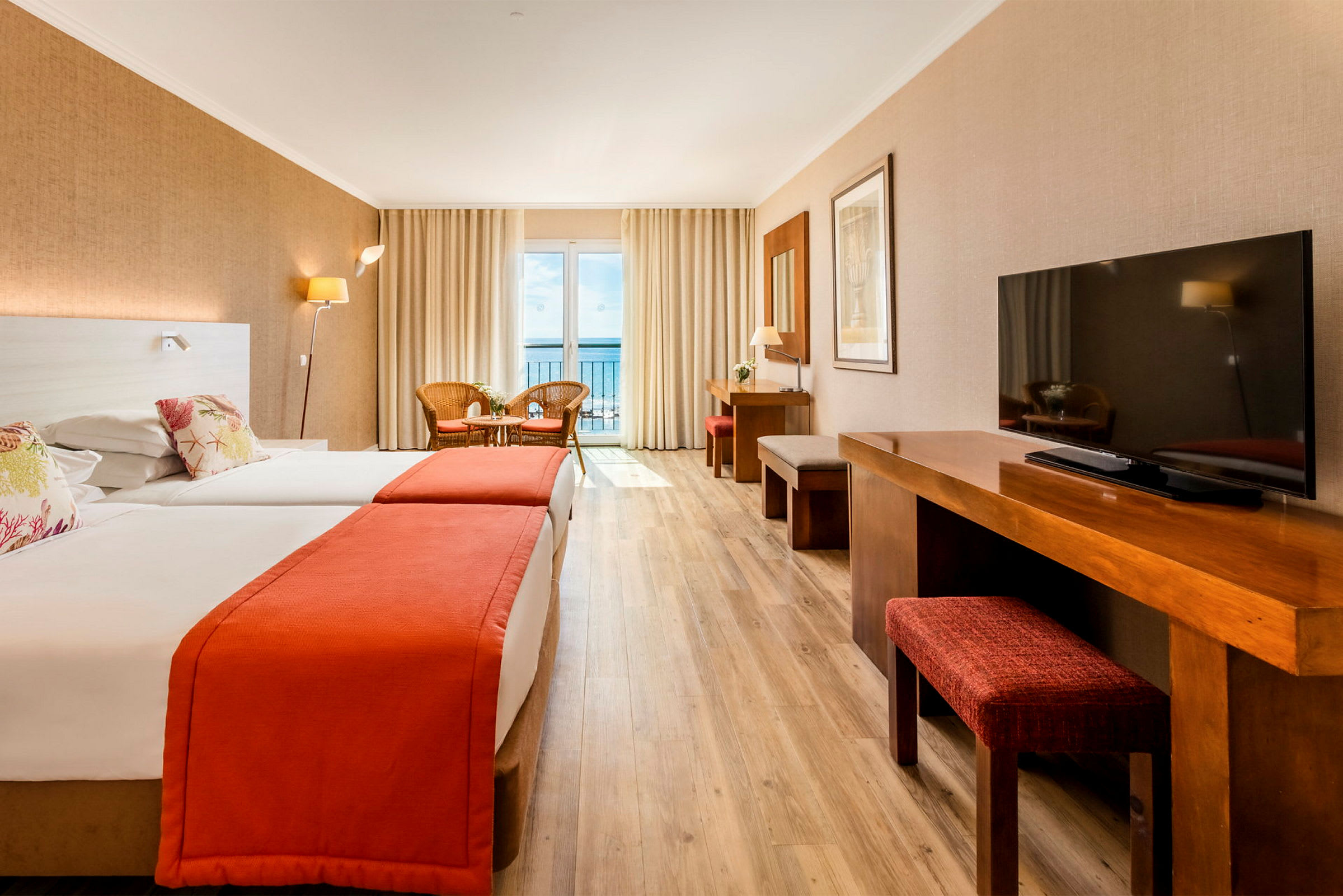 Ett hotellrum med två sängar, träbord och en TV, gardiner och en balkong med utsikt mot havet. (AI-genererad text)