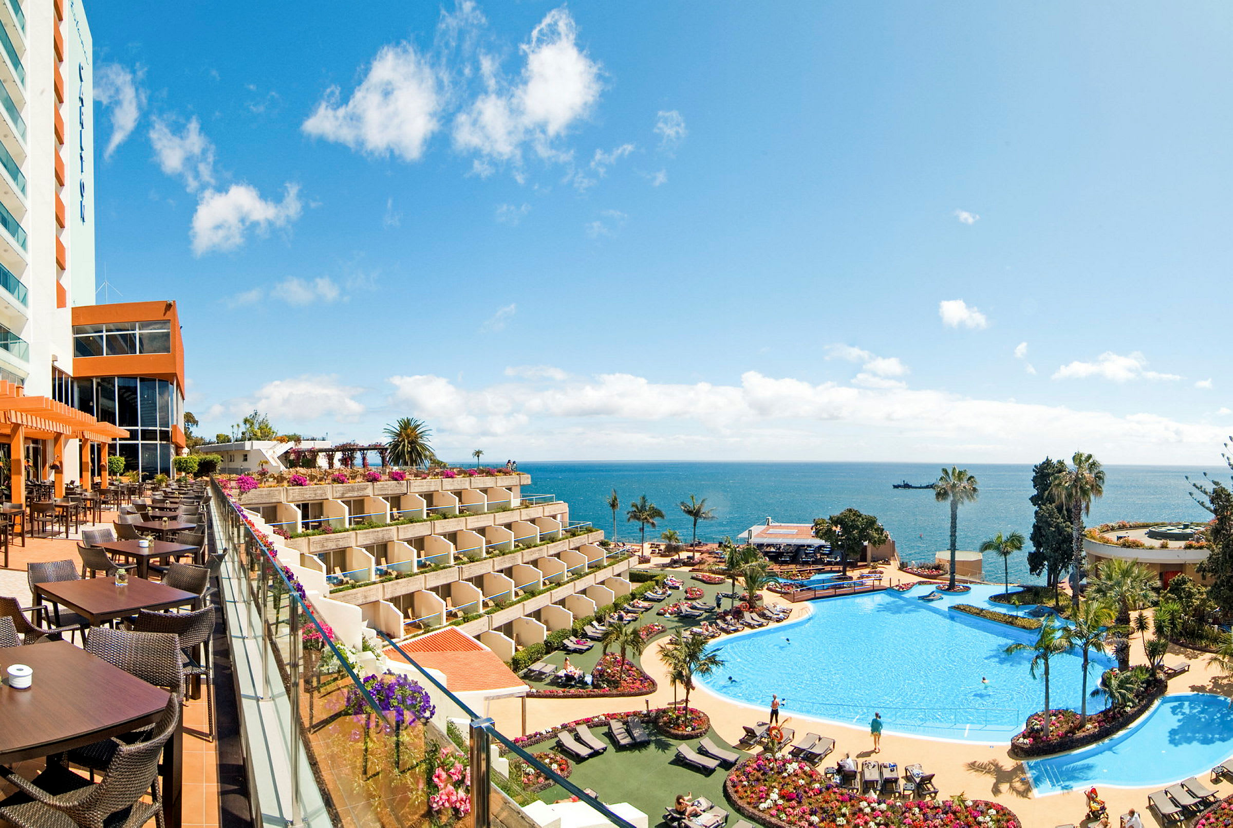 Bild av ett hotell med utsikt över havet. Det finns en stor pool omgivet av solstolar och växtlighet samt en uteservering. (AI-genererad text)