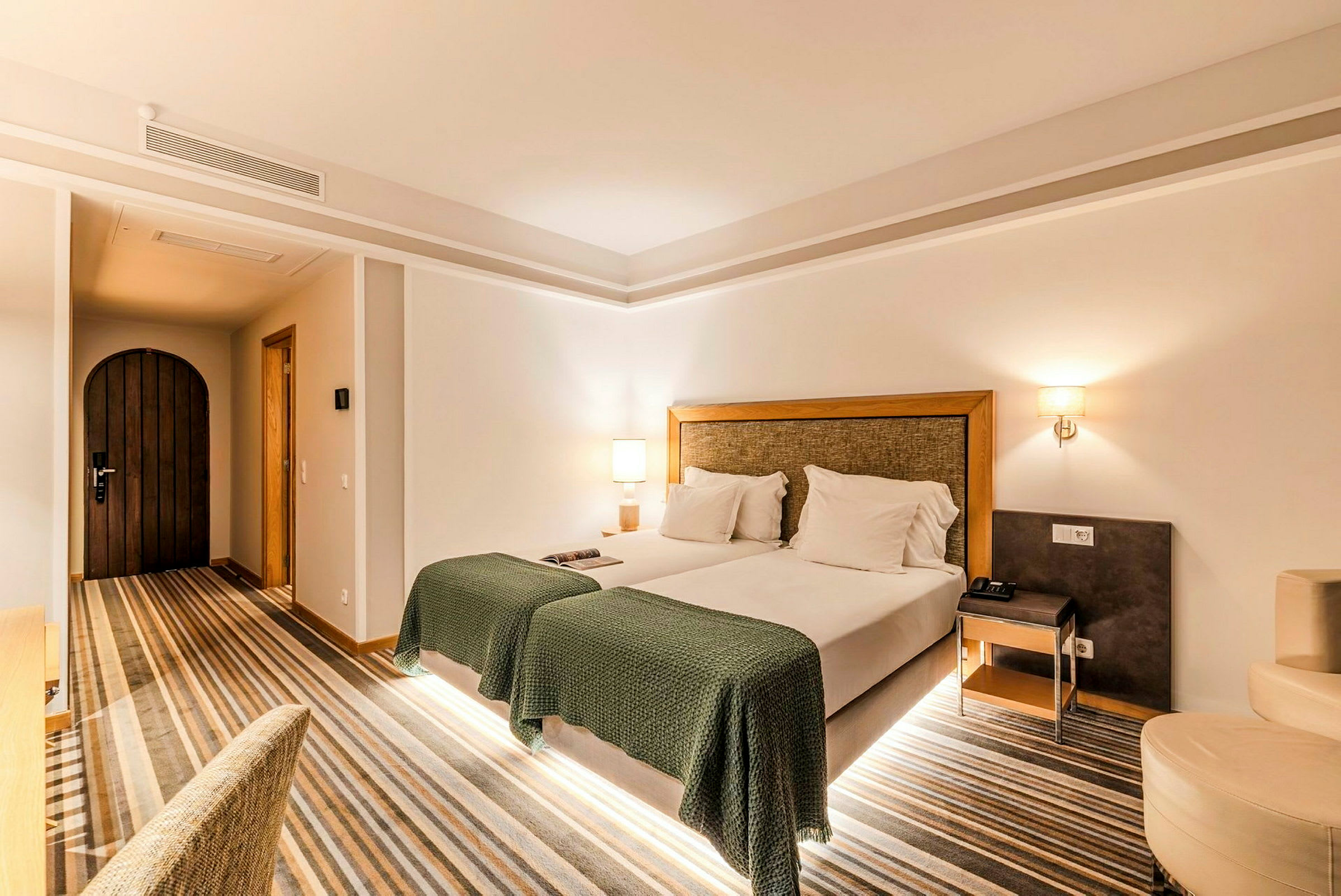 Ett hotellrum med en dubbelsäng, gröna överkast och två vita lampor på nattygsbord vid varje sida. Ett trädörr syns på ena väggen. (AI-genererad text)