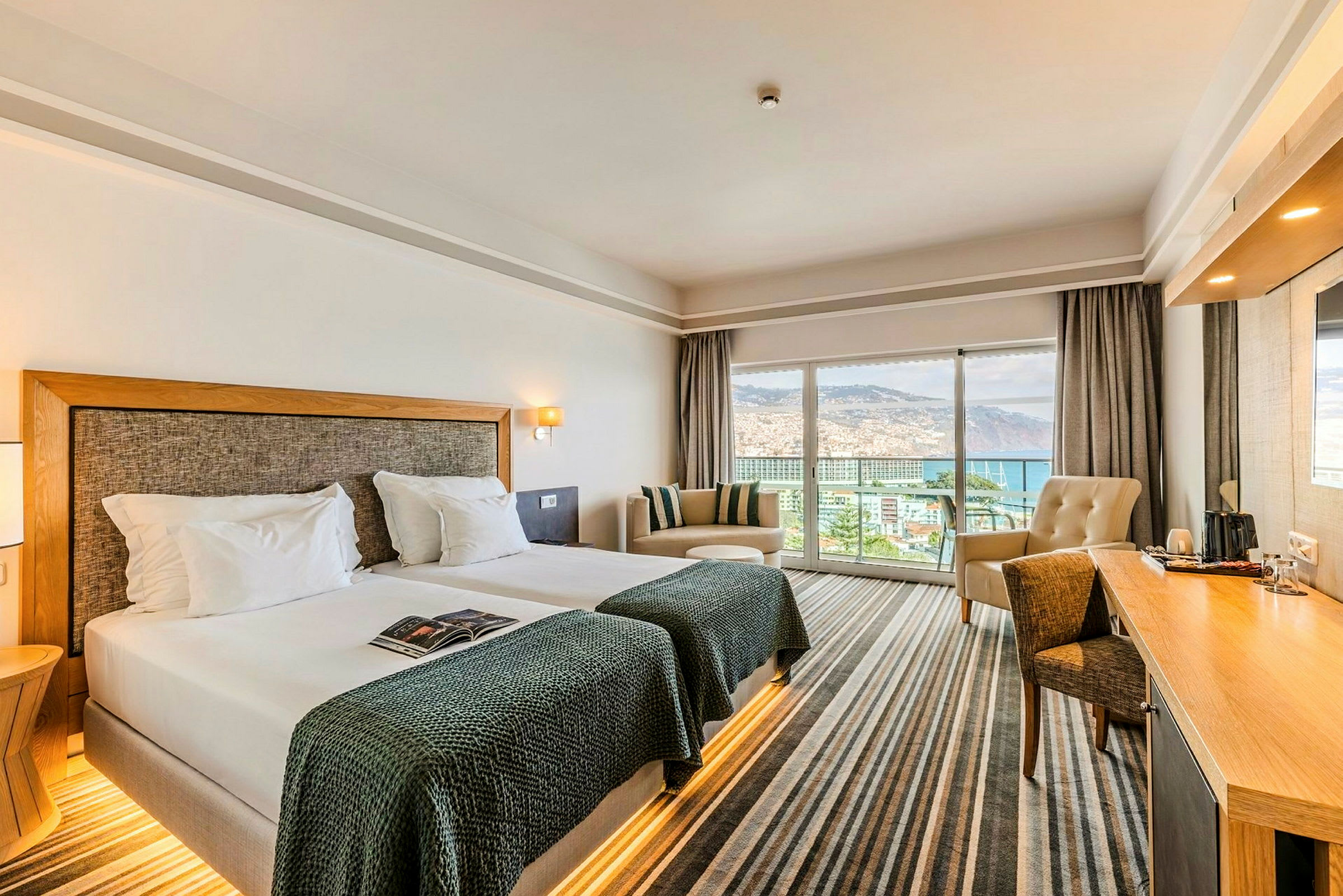 Hotellrum med två enkelsängar, en sittgrupp och ett skrivbord. Stora fönster med utsikt över havet och omgivningen. (AI-genererad text)