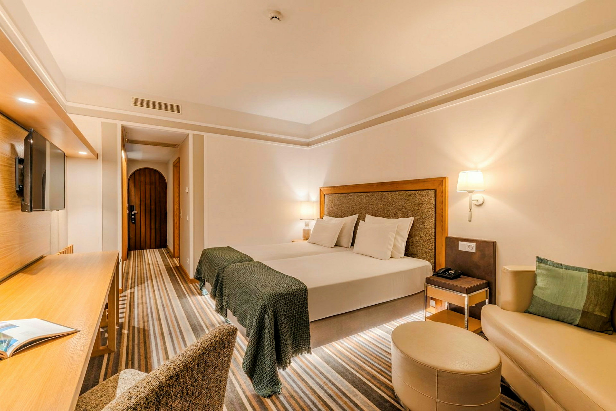 Ett välbelyst hotellrum med en stor dubbelsäng, skrivbord, stol, väggmonterad TV och en liten sittgrupp med en soffa och fotpall. (AI-genererad text)