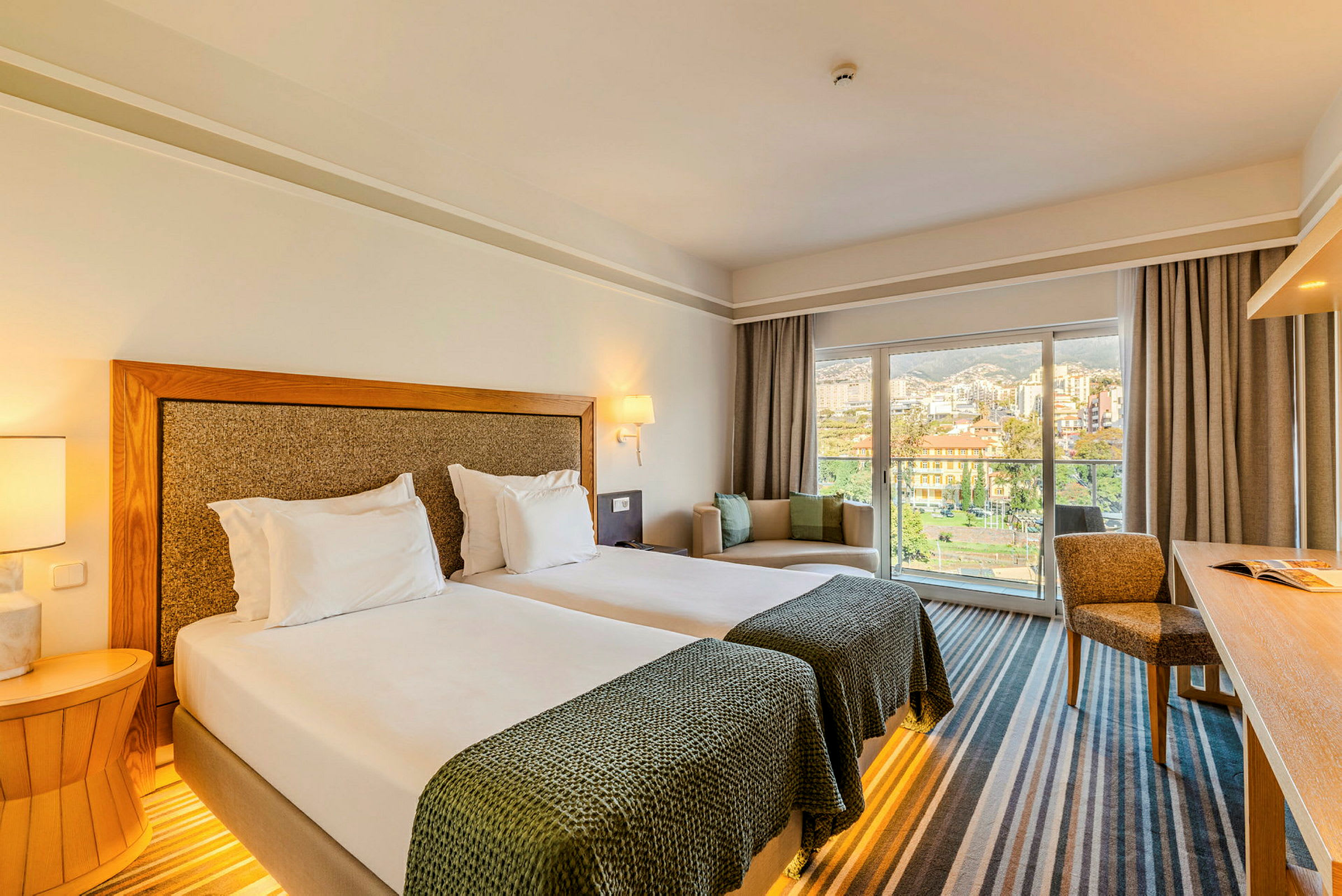 Hotellrum med två enkelsängar, sittgrupp och ett skrivbord, med stora fönster som ger utsikt mot staden. (AI-genererad text)