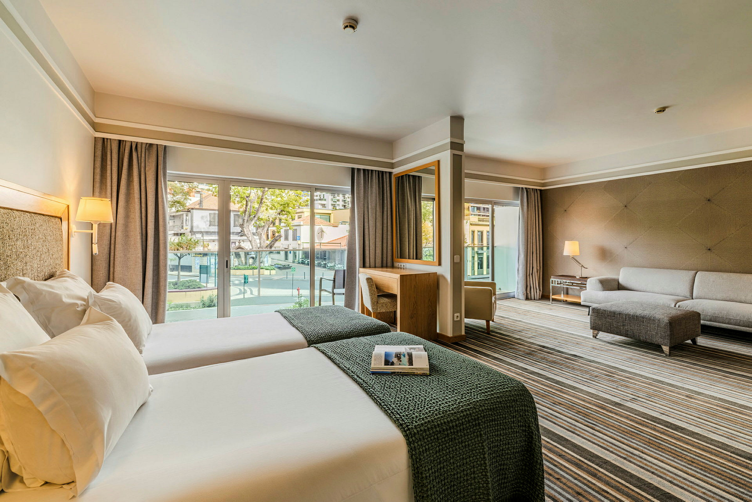 Ett hotellrum med två enkelsängar, en soffa och ett skrivbord samt fönster som leder ut till en balkong. (AI-genererad text)