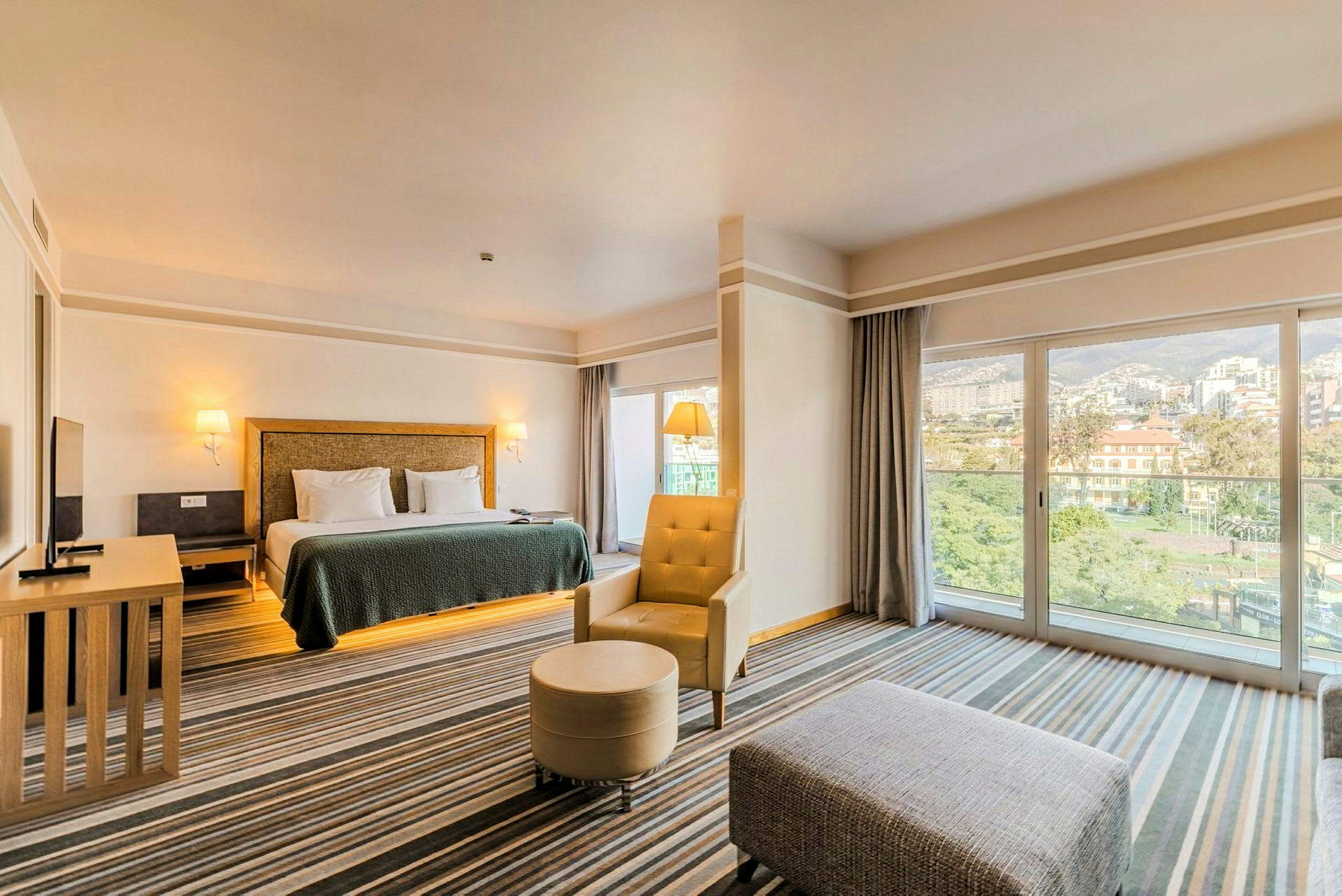 Ett hotellrum med en stor säng, fåtölj och soffbord samt stora fönster som vetter mot en stadsvy. (AI-genererad text)