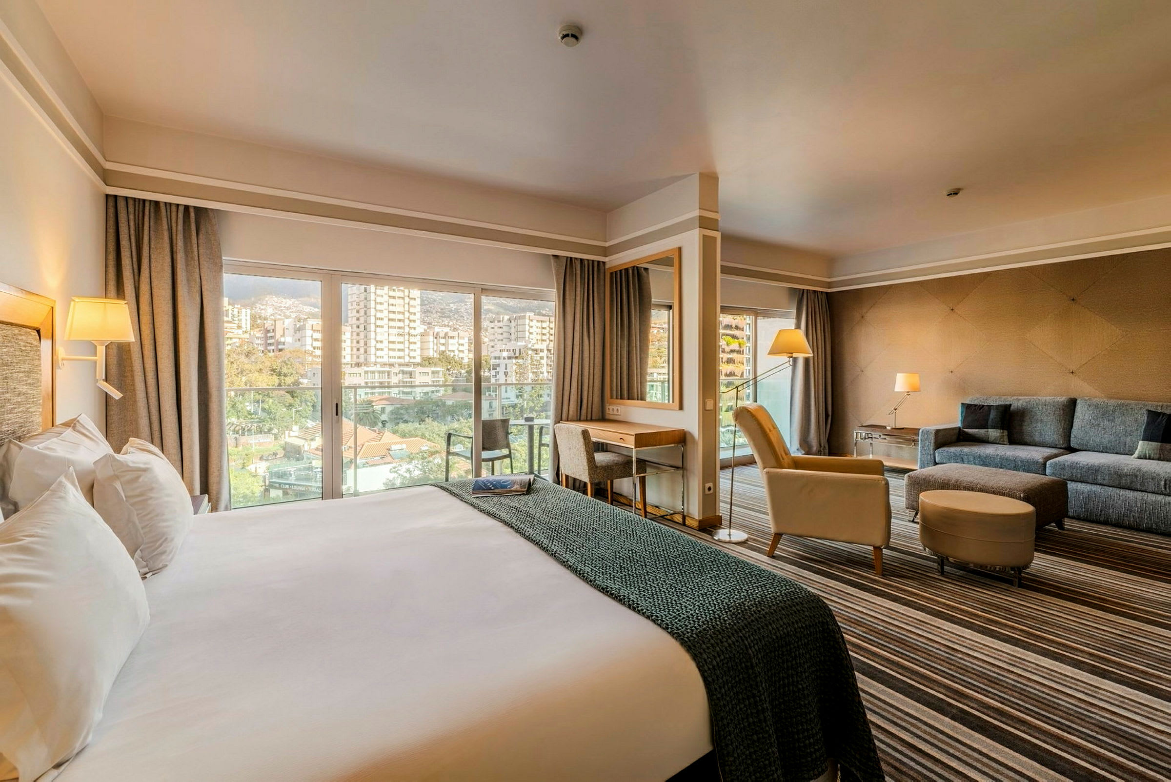 Ett hotellrum med en stor säng, sittgrupp och skrivbord. Rummet har stora fönster med utsikt över en stad. (AI-genererad text)