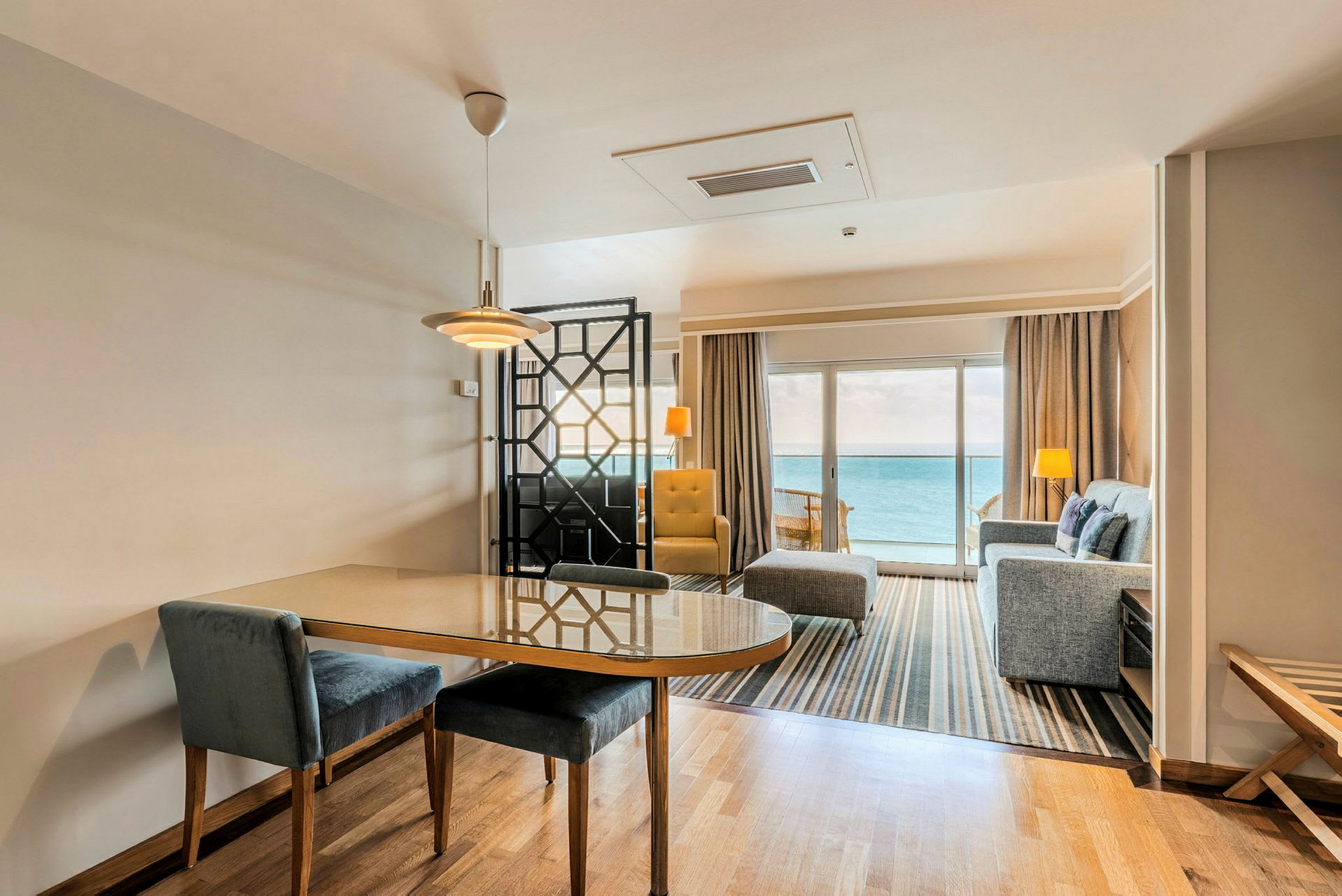Ett hotellrum med sittgrupp och matbord, med utsikt över havet från en stor glasdörr. (AI-genererad text)
