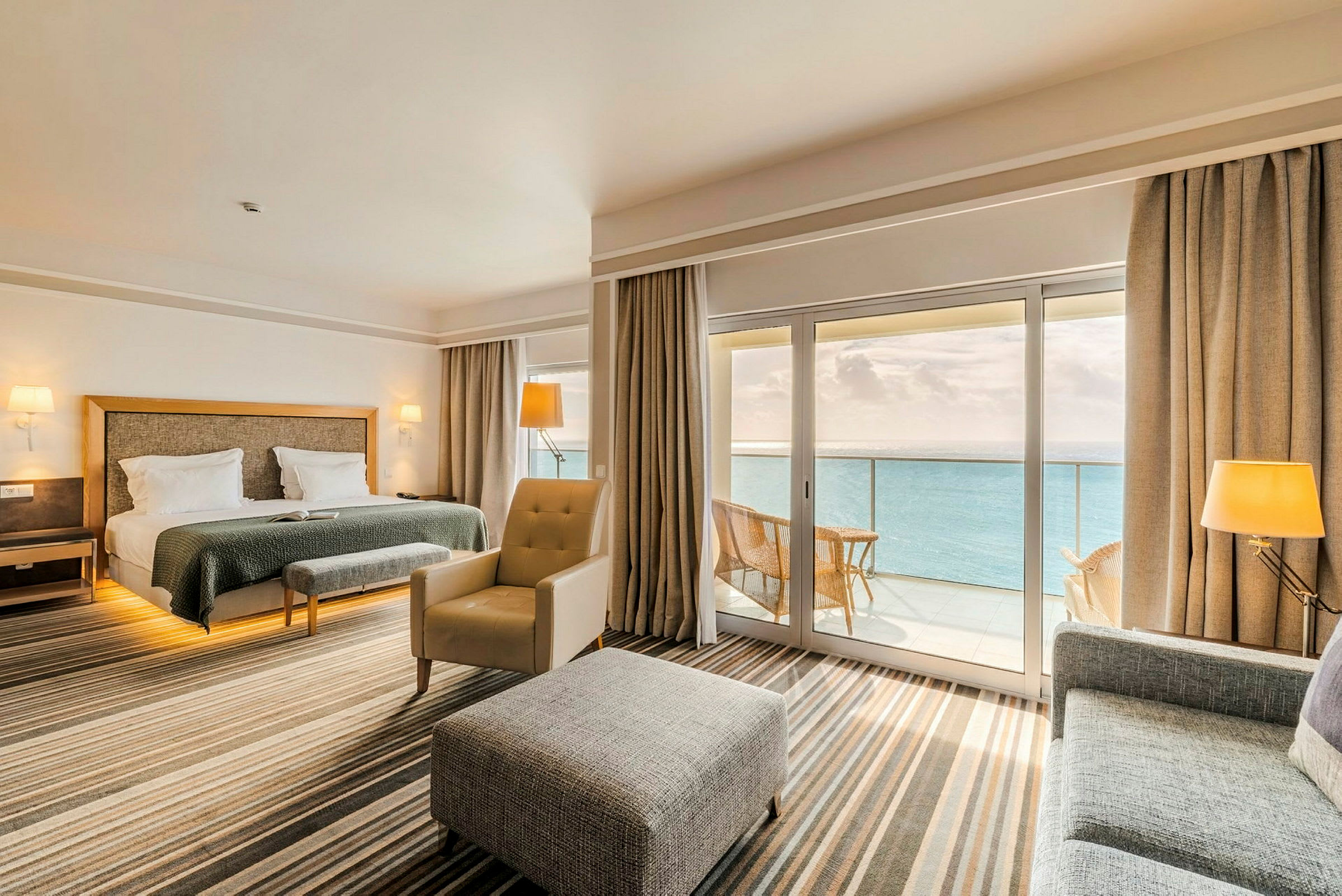 Ett hotellrum med dubbelsäng, sittgrupp och stora fönster som vetter mot en balkong med utsikt över havet. Rummet har en stilren och modern inredning. (AI-genererad text)