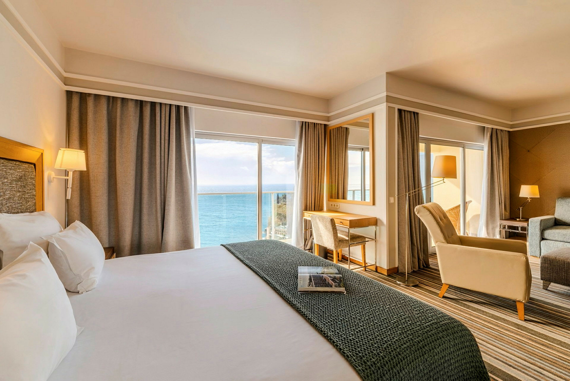 Ett hotellrum med stor säng, sittgrupp och skrivbord, med utsikt över havet genom stora fönster. (AI-genererad text)