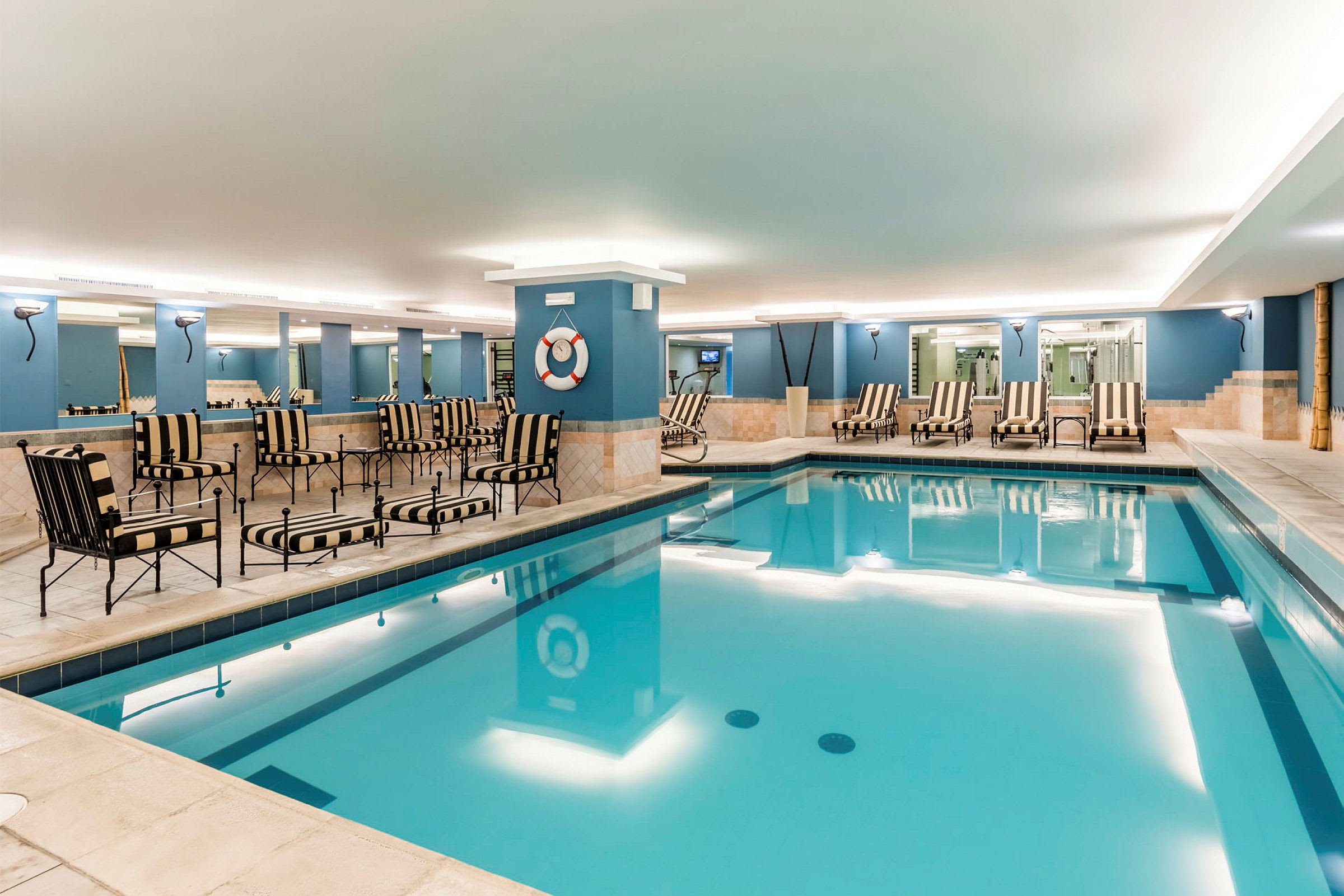 En inomhuspool med blått vatten, omgiven av bord och stolar med randiga mönster samt solstolar. (AI-genererad text)