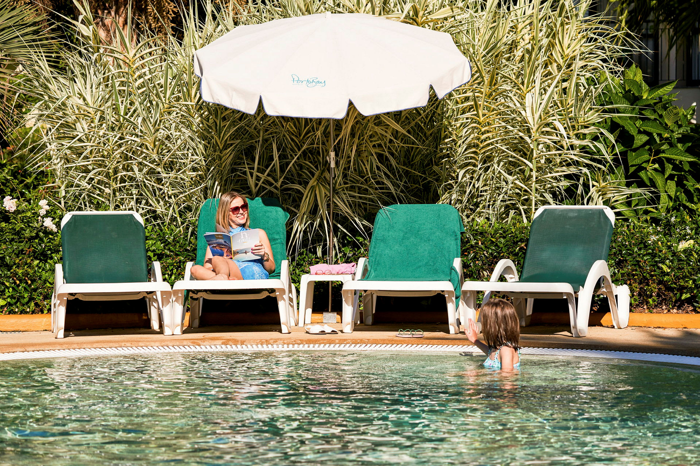 Två solstolar vid en pool, en person sitter och läser under ett parasoll medan en annan är i poolen. (AI-genererad text)