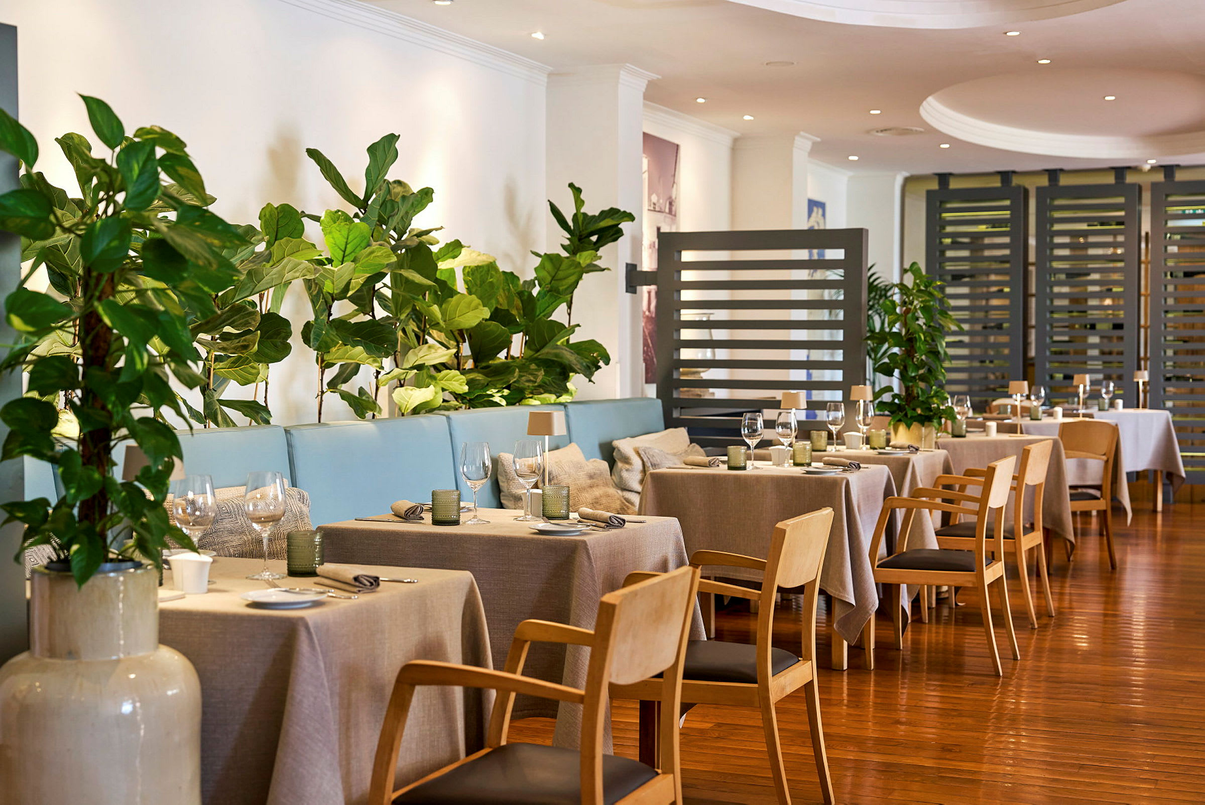 Ett välskött inomhusområde av en restaurang med eleganta trämöbler, dukade bord, frodiga gröna växter, och ljusa väggar. (AI-genererad text)