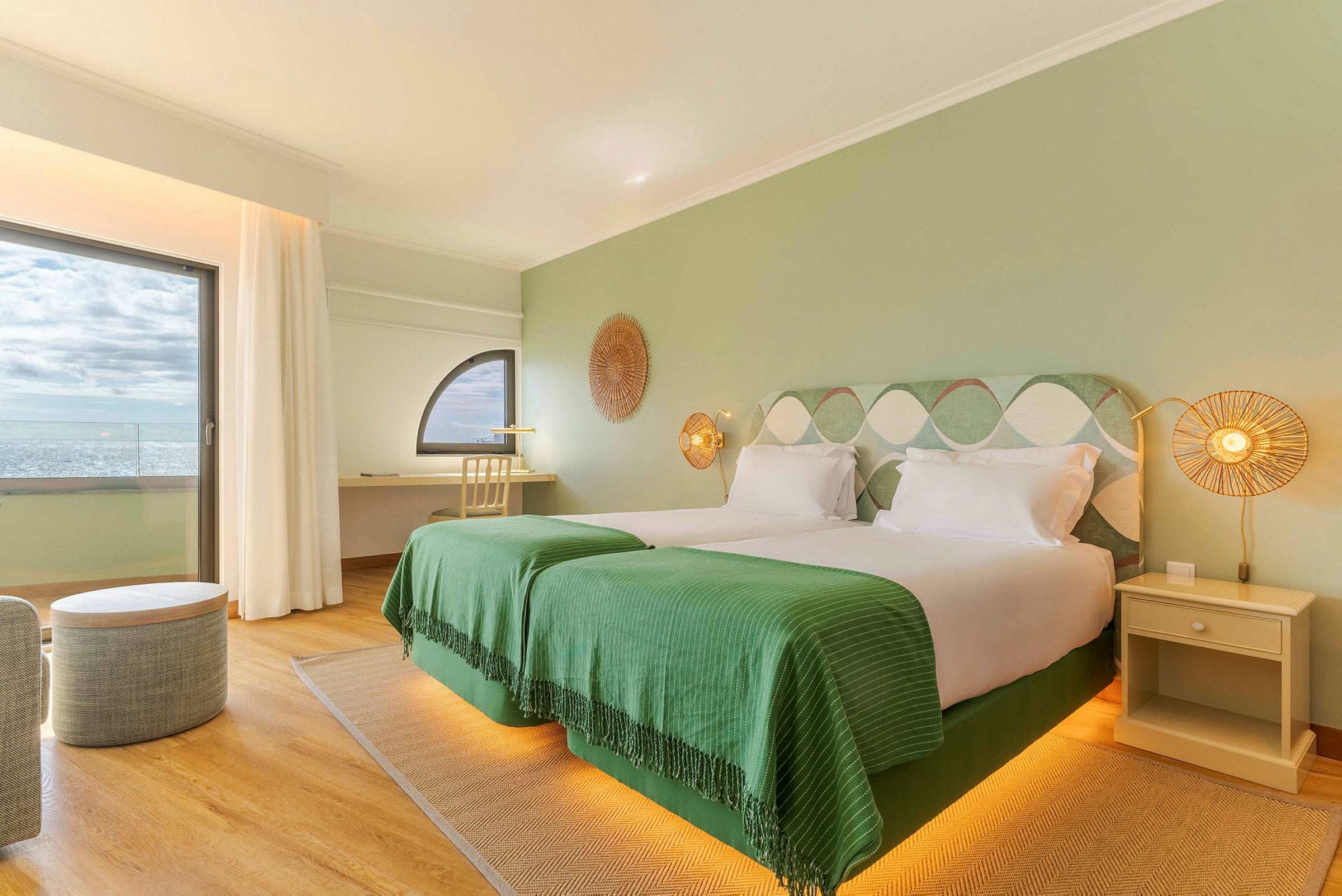 Ett modernt hotellrum med två enkelsängar täckta med gröna filtar, ett fönster med havsutsikt och stilrena inredningsdetaljer. (AI-genererad text)