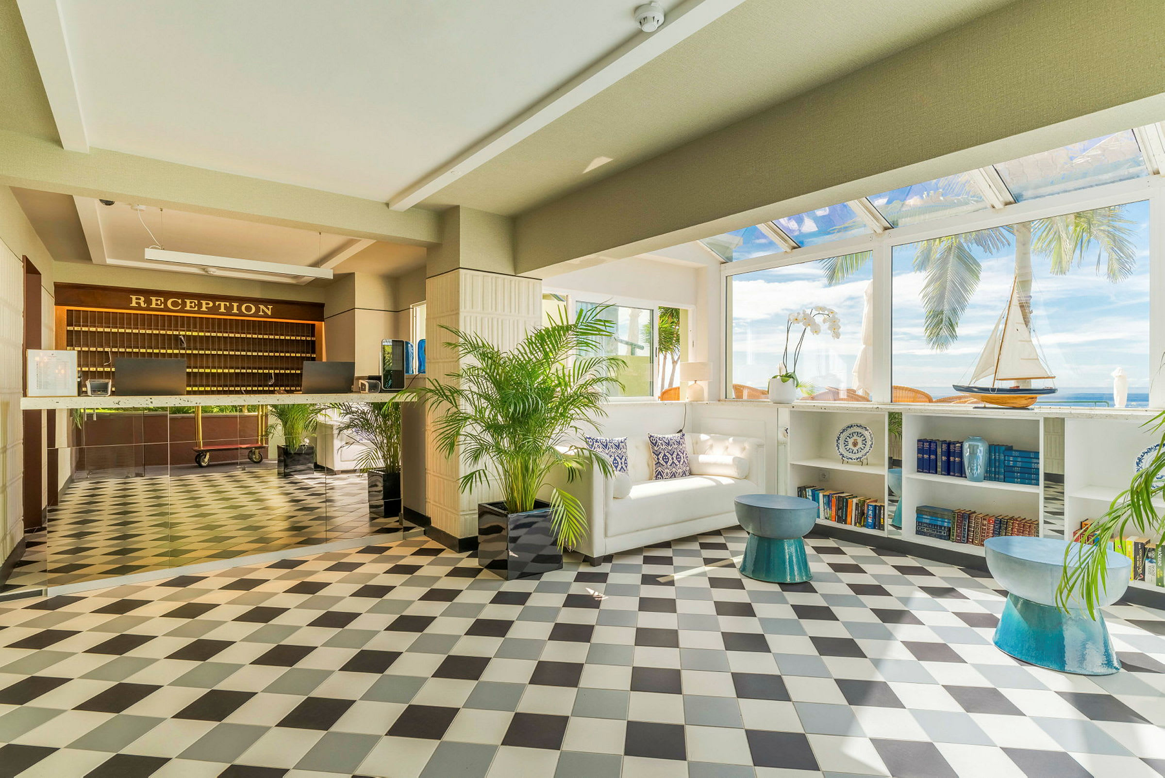 Hotellreception med svartvitt golv, en välkomnande soffa och dekorativa element som en modellsegelbåt och böcker i hyllan samt utsikt över palmer och havet genom breda fönster. (AI-genererad text)