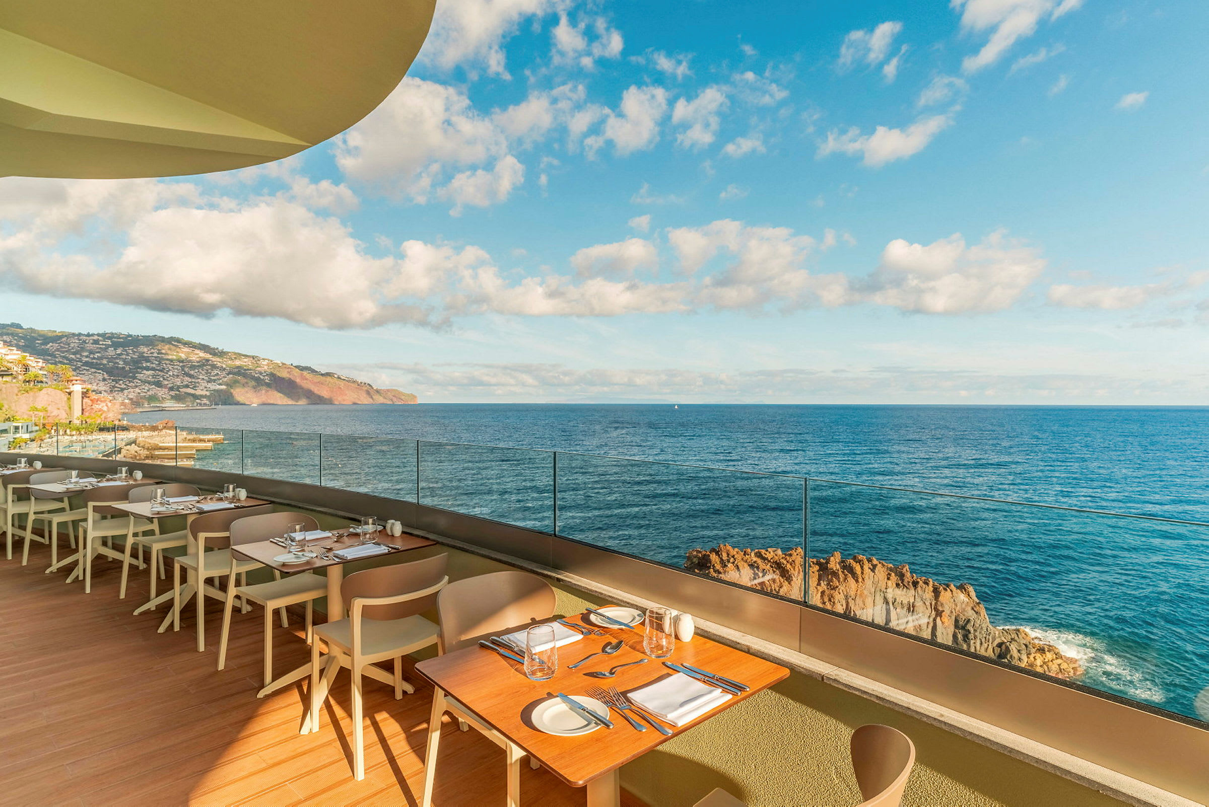 Restaurangterrass med bord och stolar, med utsikt över havet och en klippig kustlinje under en klar himmel. (AI-genererad text)