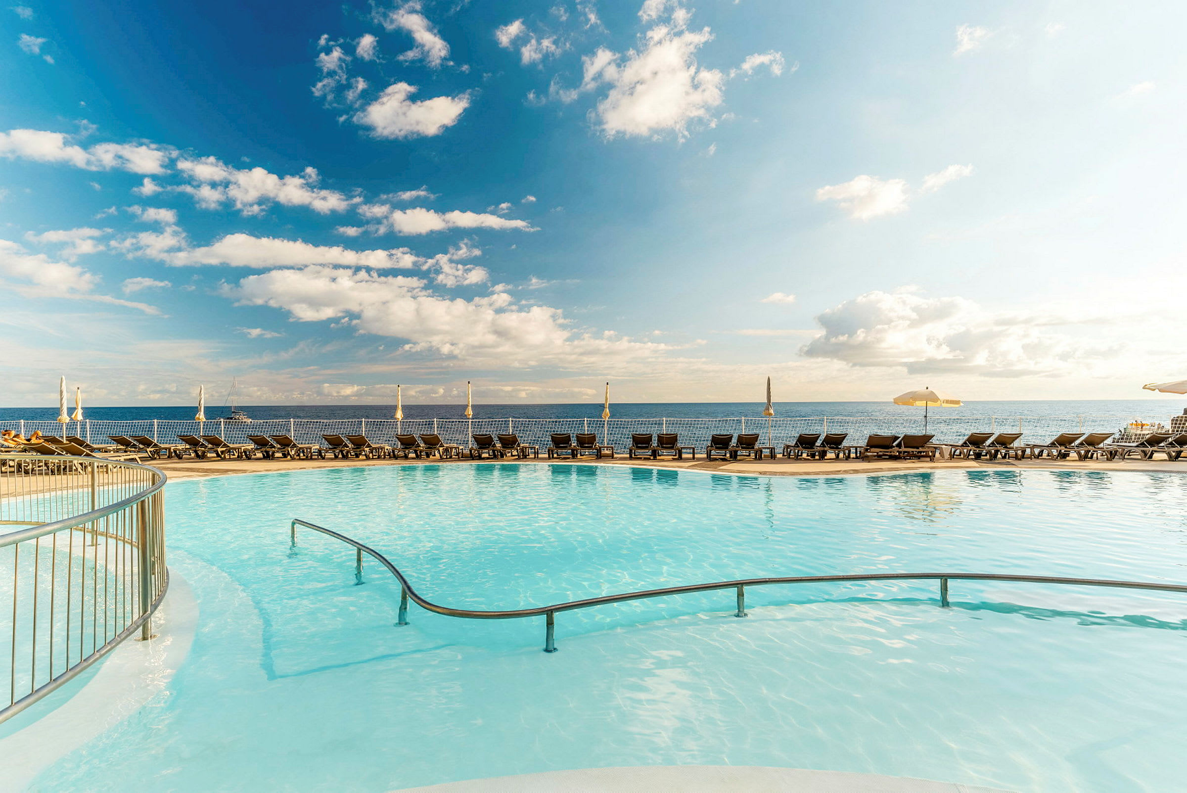 En pool med klart vatten, omgiven av solstolar och parasoller, med havet och himlen i bakgrunden. (AI-genererad text)