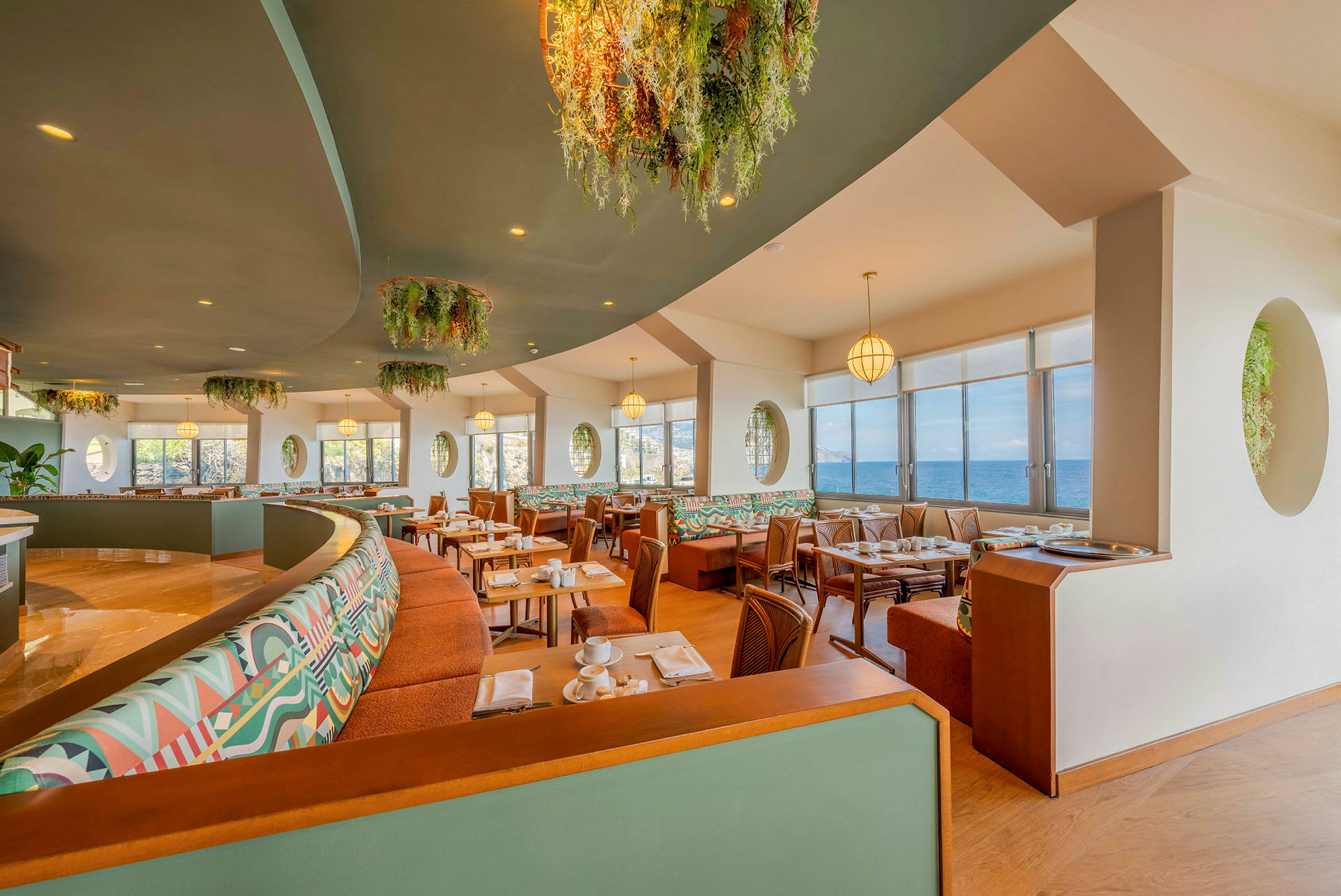 Restaurang med stora fönster och utsikt över havet, inredd med färgglada soffor och gröna dekorationer. (AI-genererad text)