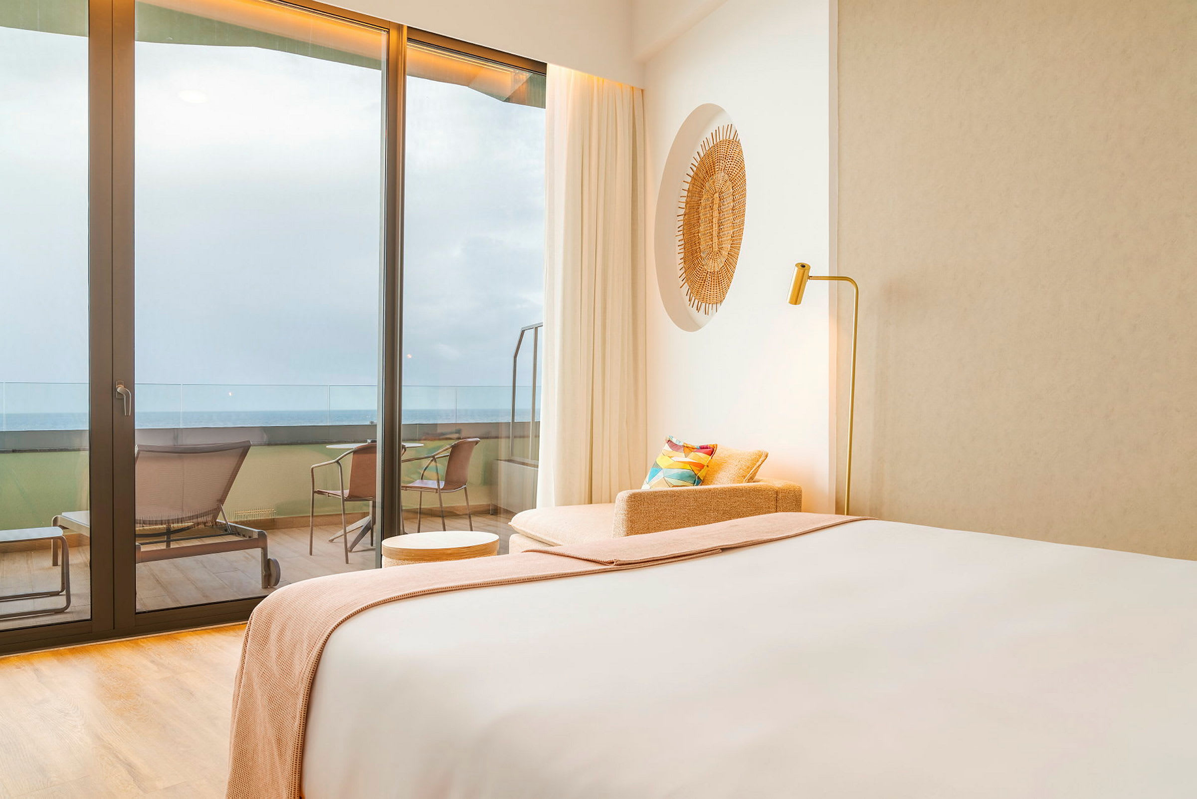 Ett hotellrum med modern inredning. Det finns en säng, en fåtölj och ett litet bord närmast en glasyta med en utsikt över en balkong och havet. (AI-genererad text)