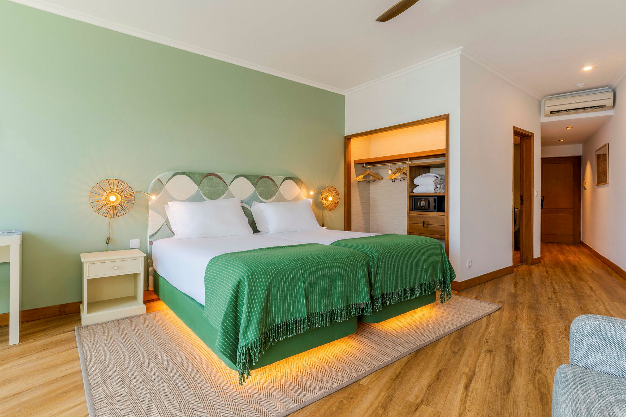 Ett hotellrum med två enkelsängar klädda med gröna överkast, belysta från undersidan. Rummet har trägolv, en garderob och vägg i grön nyans. (AI-genererad text)