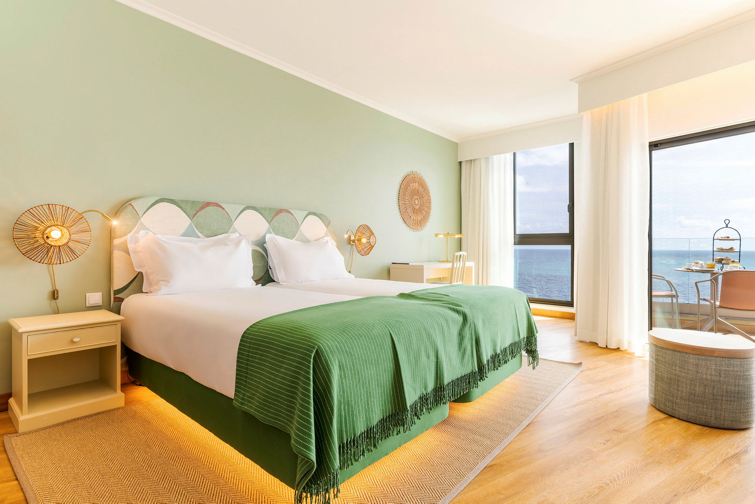 Ett ljust hotellrum med dubbelsäng, grön inredning och stora fönster som erbjuder havsutsikt. (AI-genererad text)
