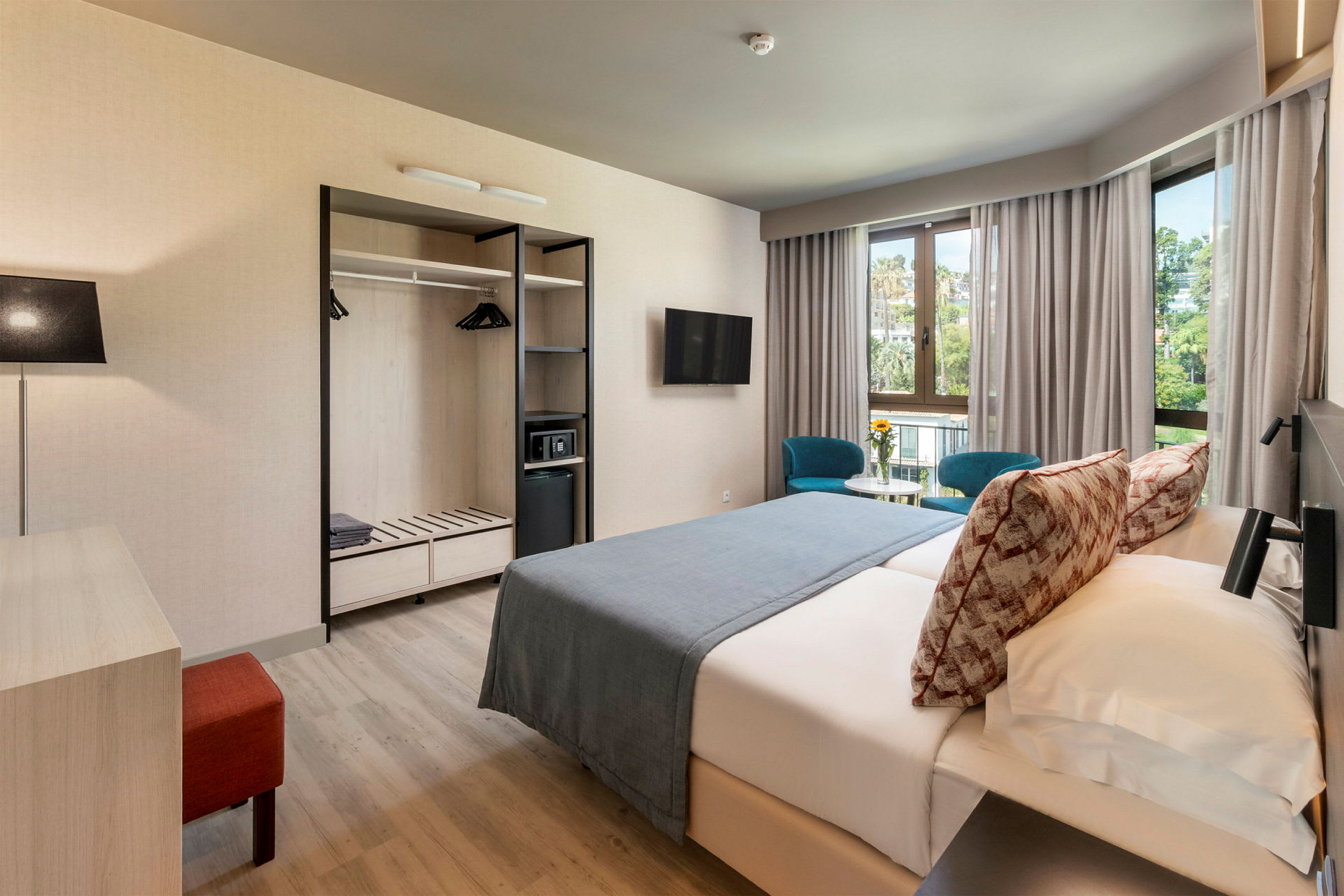 Ett hotellrum med dubbelsäng, två blå stolar vid ett bord framför fönster samt garderob med öppna hyllor. (AI-genererad text)