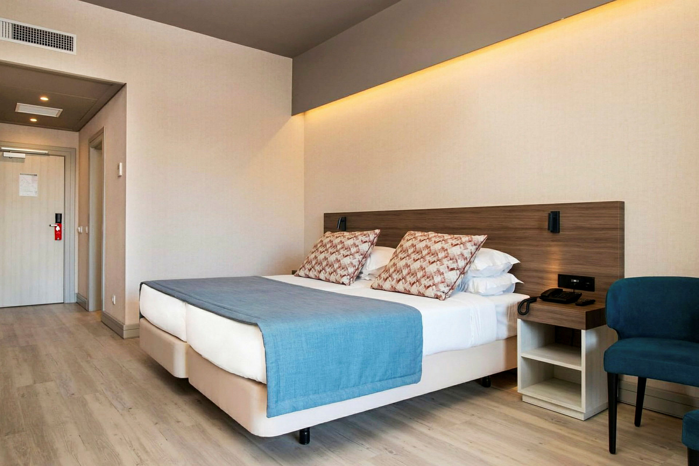 Hotellrum med en dubbelsäng, blå överkast och kuddar, vid sidan av en stol och ett nattduksbord. (AI-genererad text)
