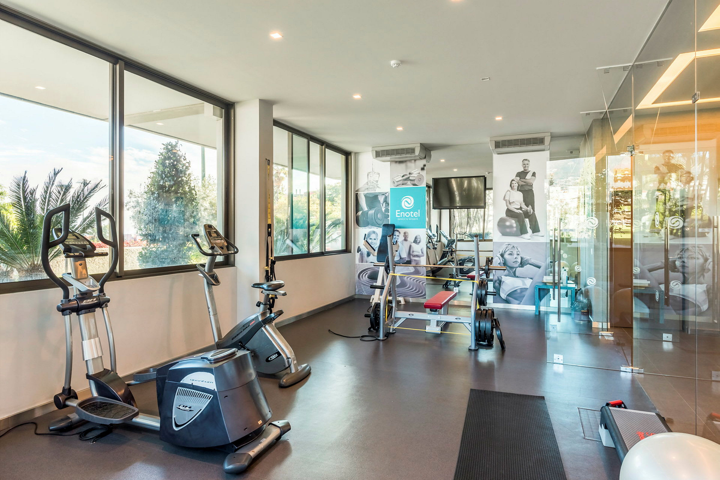 Ett gym med träningsutrustning inklusive motionscyklar och styrketräningsmaskiner, med stora fönster som släpper in naturligt ljus. (AI-genererad text)