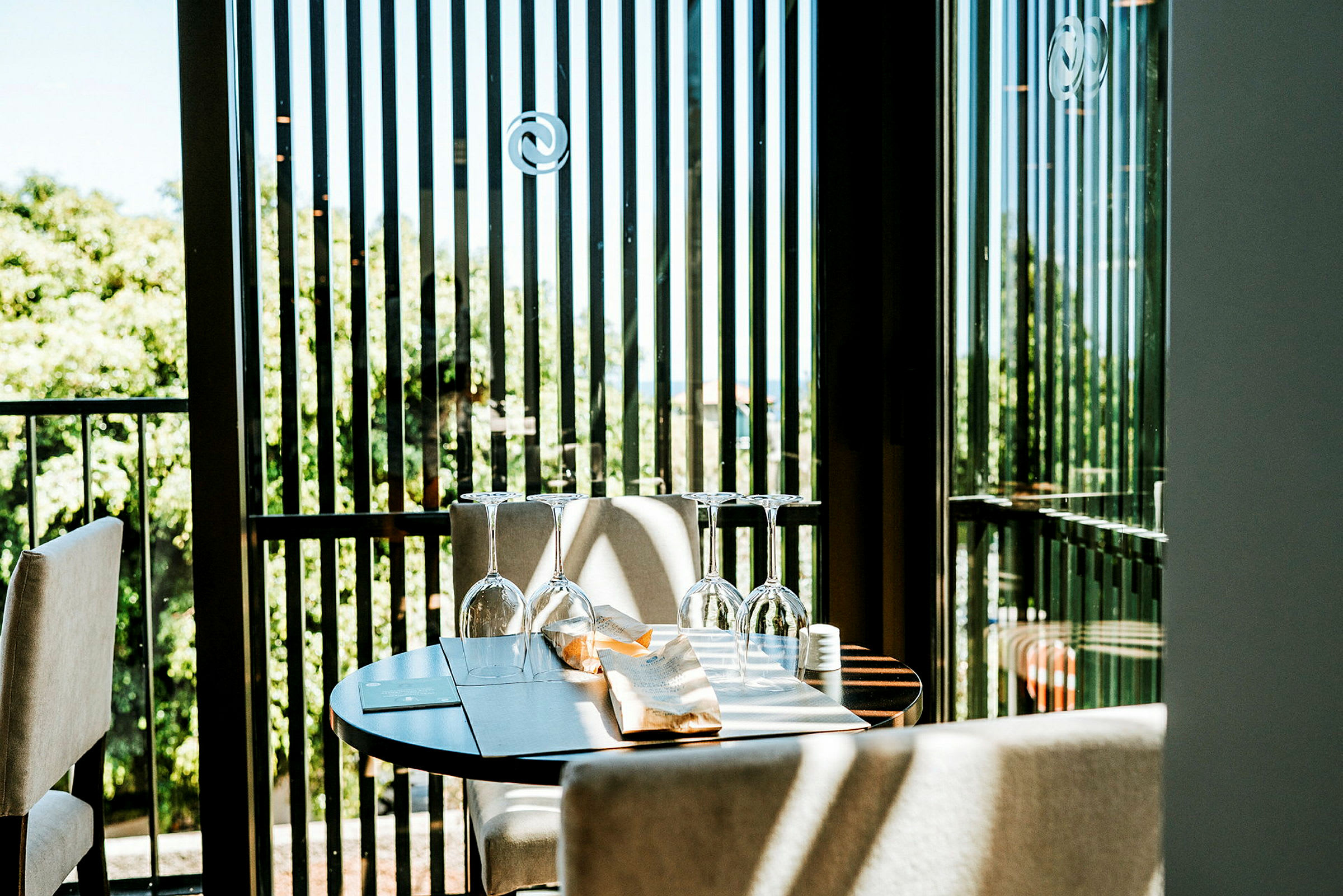 Ett dukat bord med glas på en terrass med gröna träd i bakgrunden. (AI-genererad text)