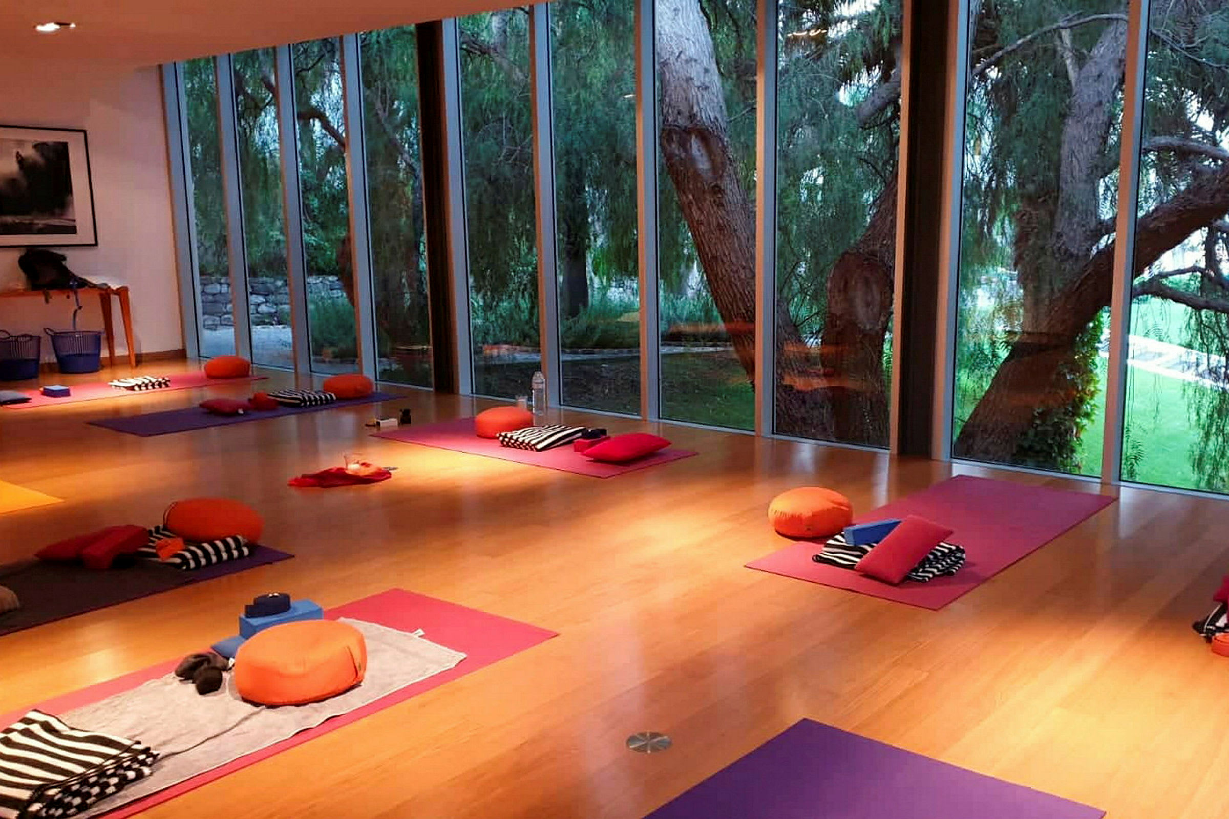 Ett rum med yoga- eller meditationsmattor, kuddar och filtar placerade på golvet, nära stora fönster som visar ett grönområde utanför. (AI-genererad text)
