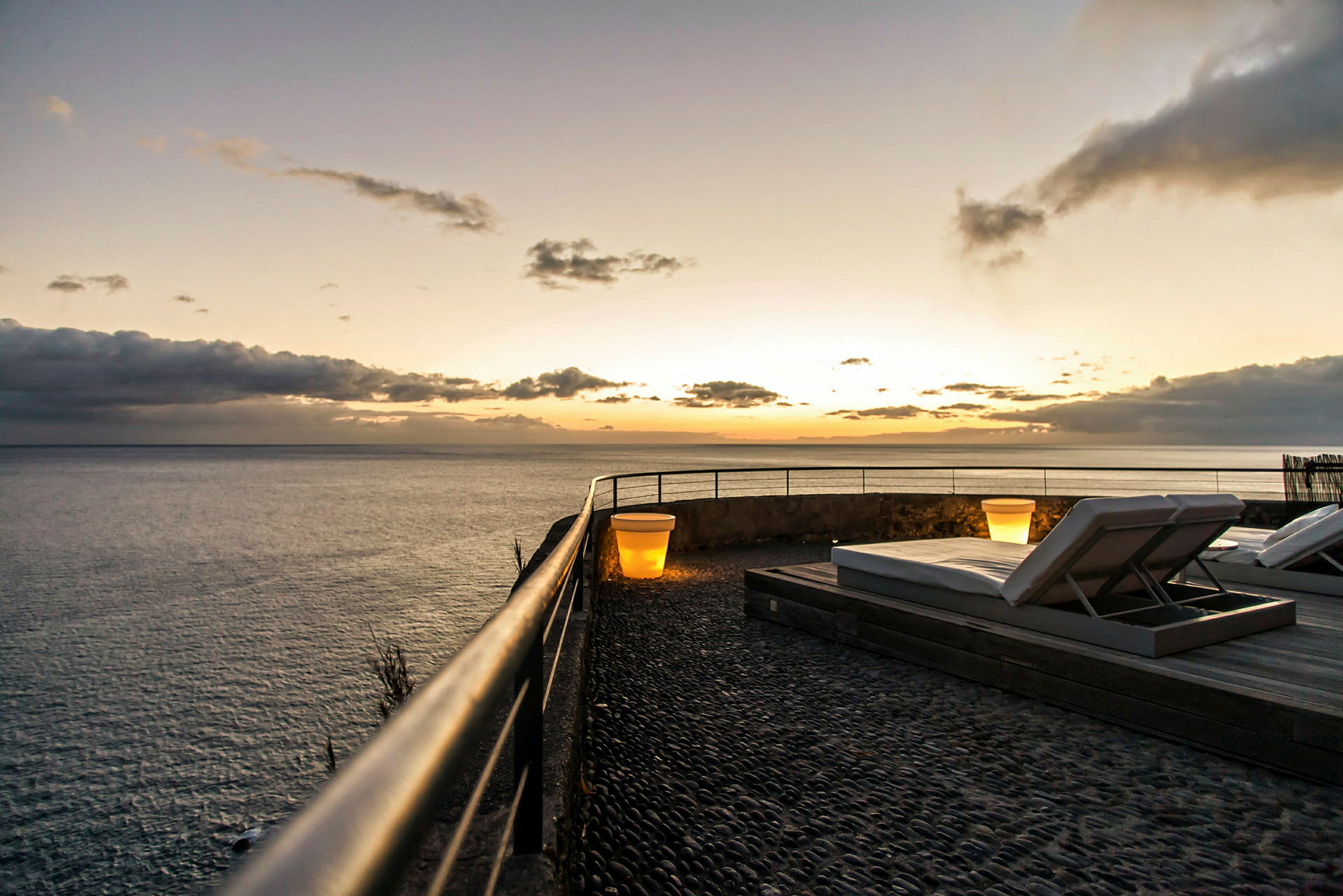 Terrass vid havet med solstolar och en lampa i solnedgång. (AI-genererad text)