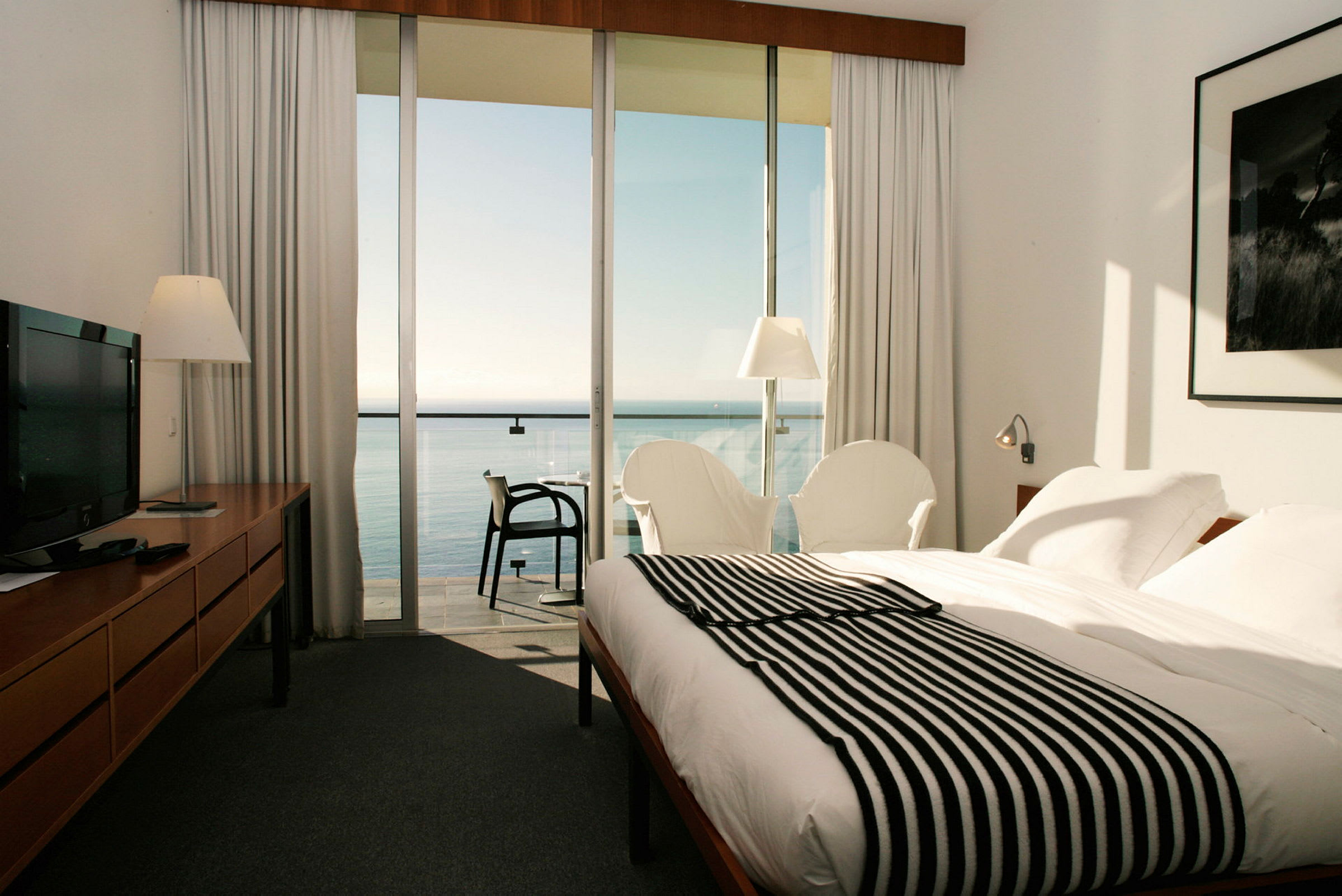 Hotelrum med säng, fåtöljer och utsikt över havet genom stora glasfönster. (AI-genererad text)