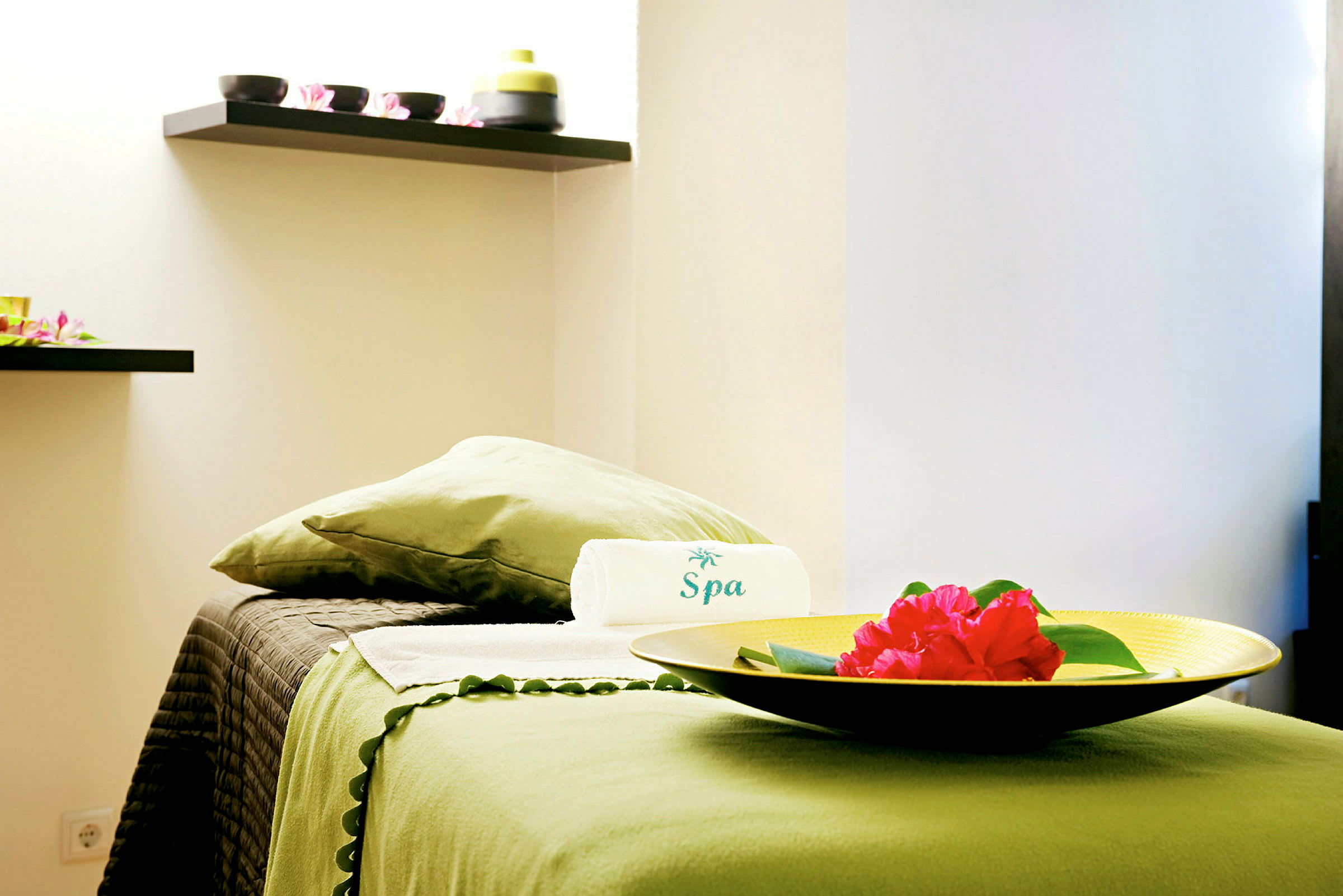 En spa-miljö med en bädd täckt av grönt tyg, en ihoprullad handduk med texten 'Spa' och en gul skål med röda blommor. (AI-genererad text)