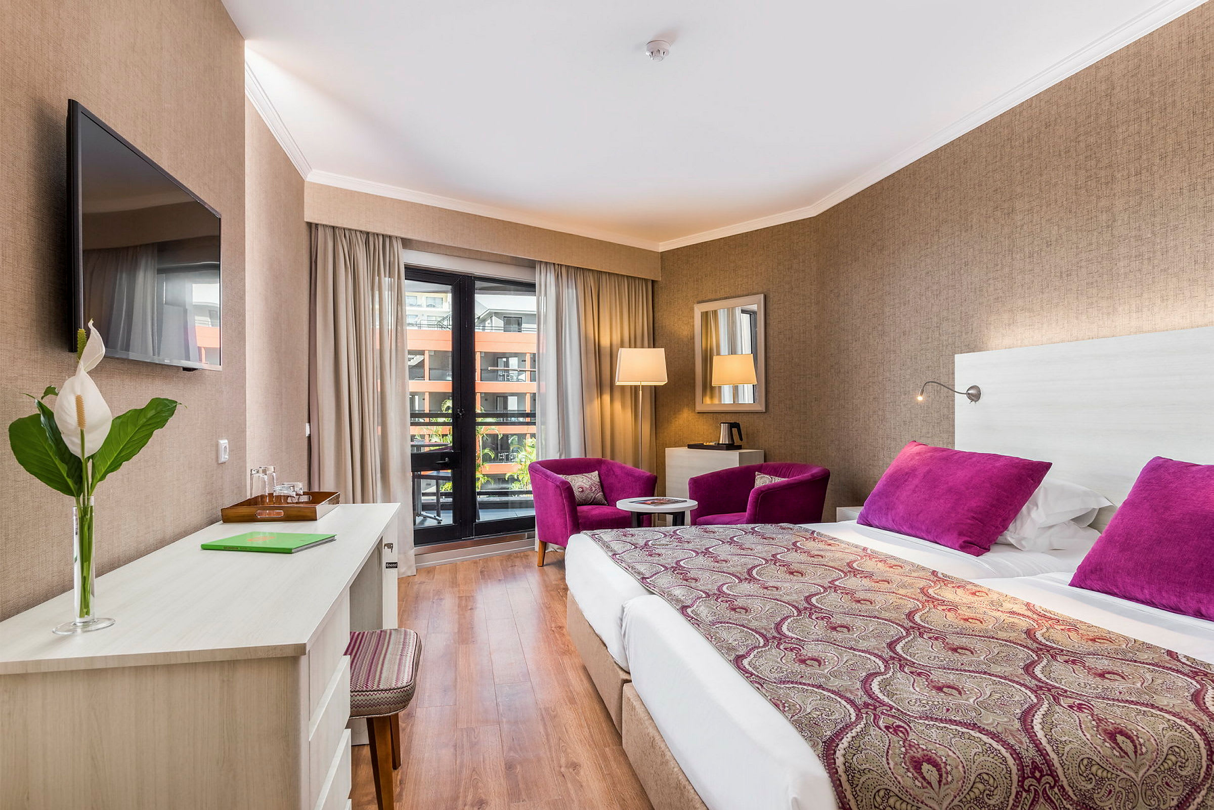 Hotellrum med dubbelsäng, skrivbord, två lila fåtöljer och balkong. (AI-genererad text)