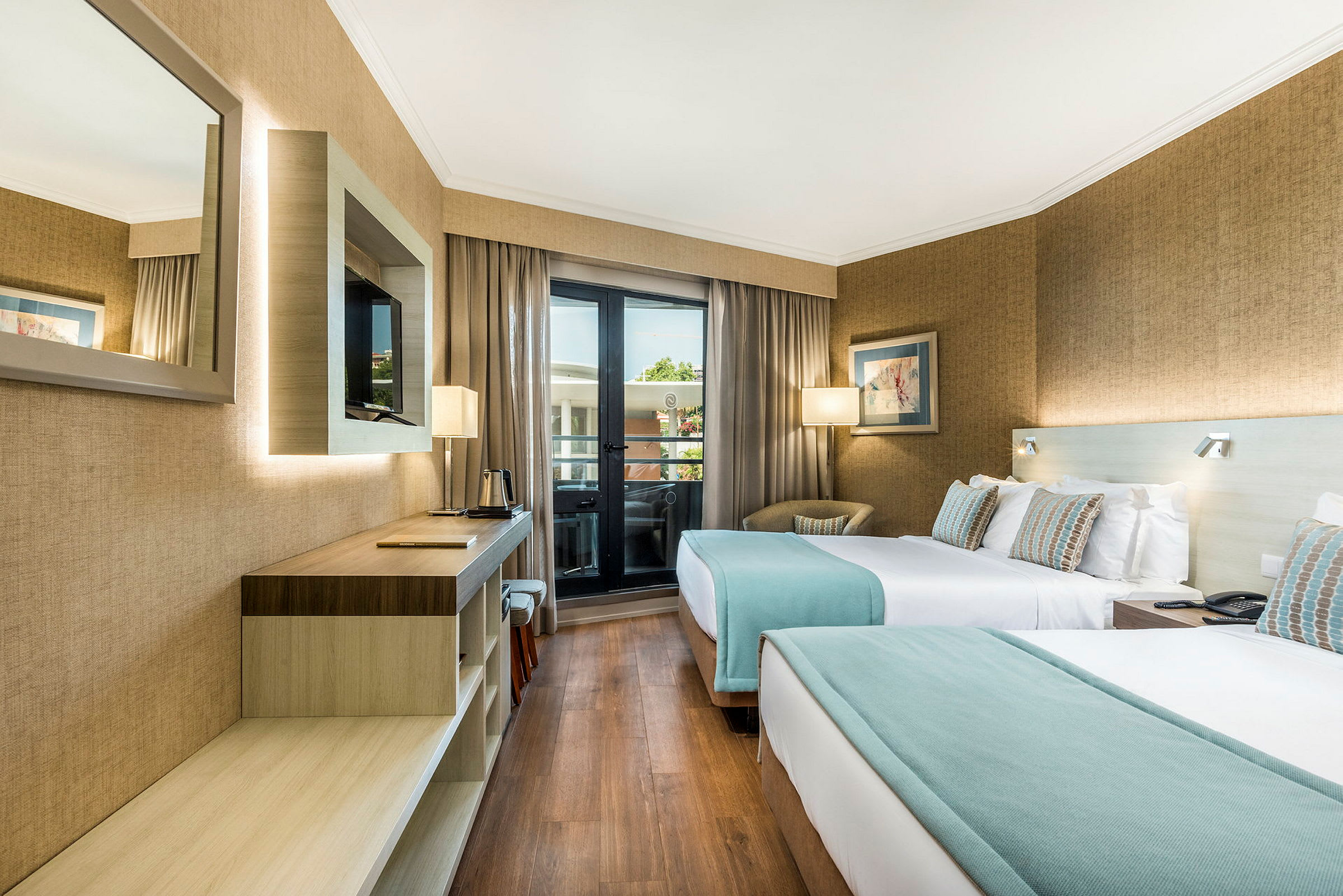 Ett hotellrum med två enkelsängar, en skrivbordsyta, en väggmonterad TV och ett fönster som leder till en balkong. (AI-genererad text)