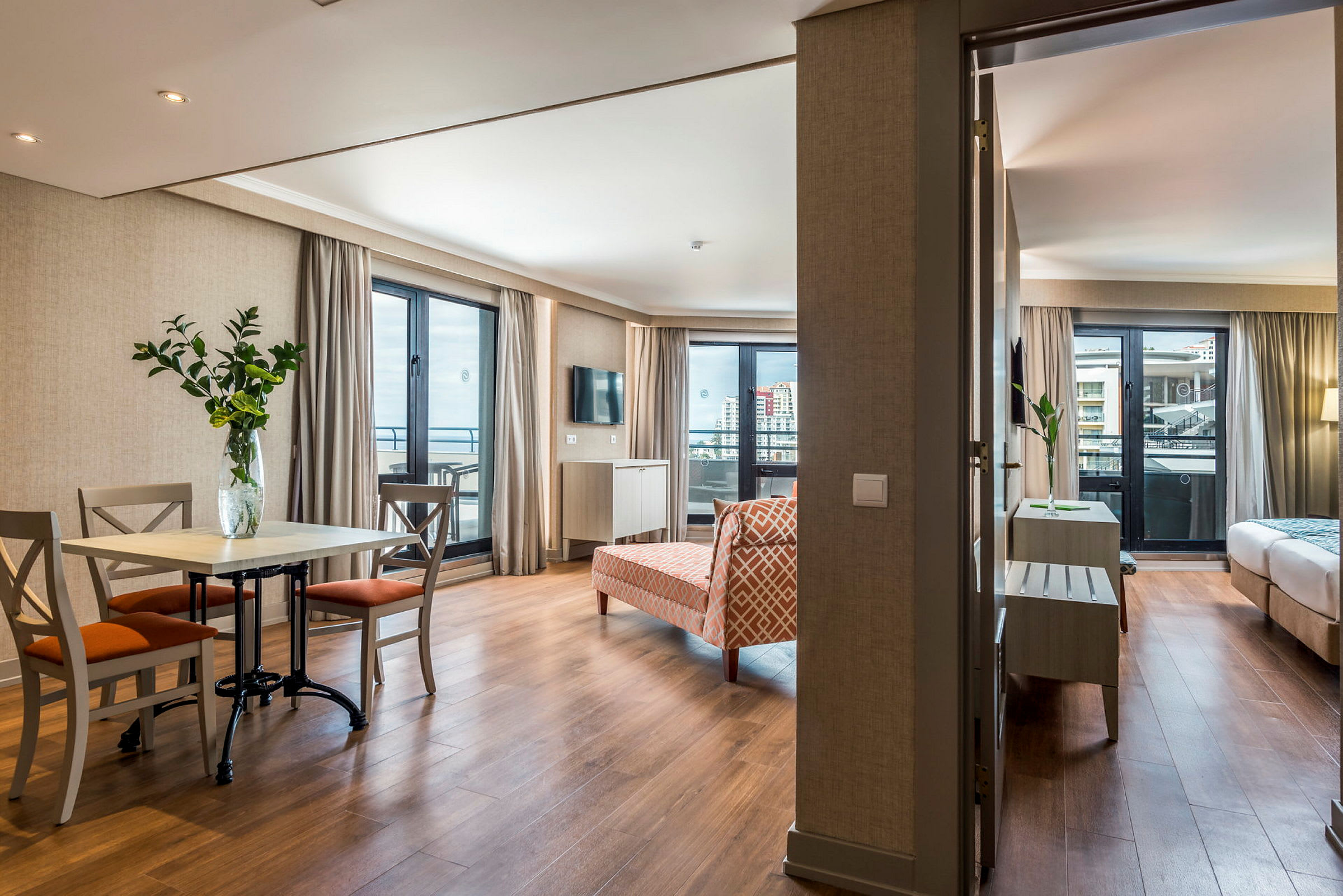 Ett hotellrum med ett vardagsrum inklusive matbord och stolar, samt ett sovrum synligt i bakgrunden. (AI-genererad text)