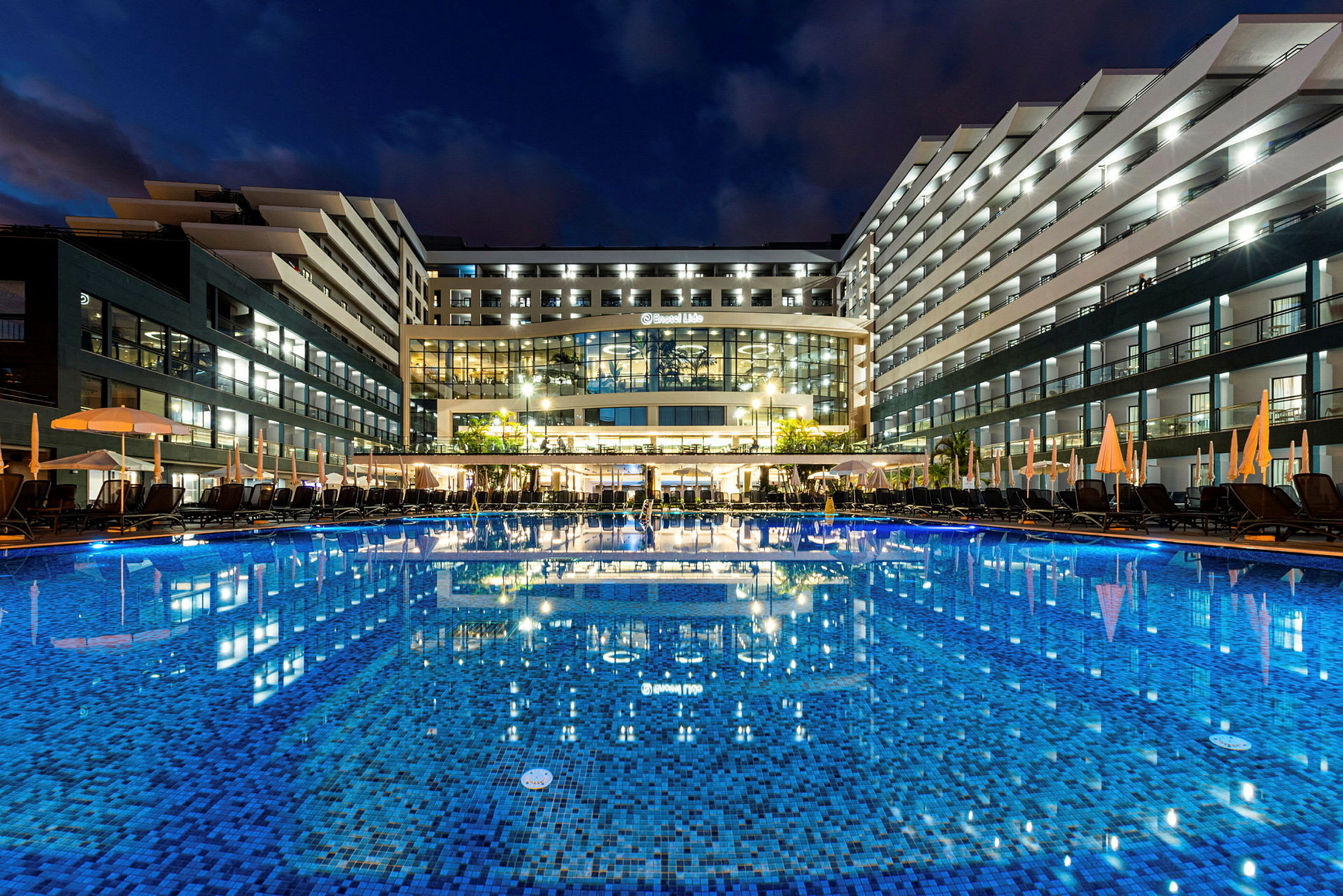 Hotell med upplysta balkonger och en stor pool framför, kvällsmiljö. (AI-genererad text)