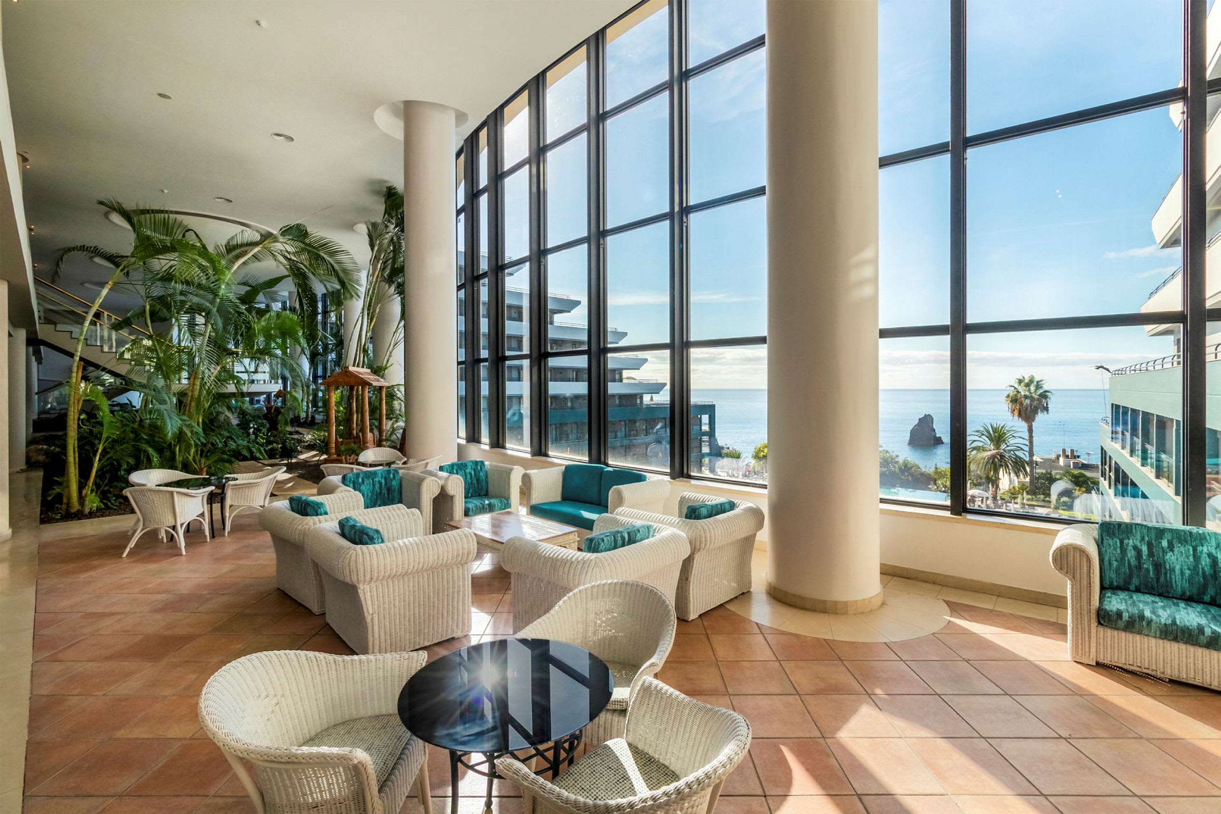 Hotellobby med stora fönster mot havet. Bord och stolar i flätade material samt växter. (AI-genererad text)