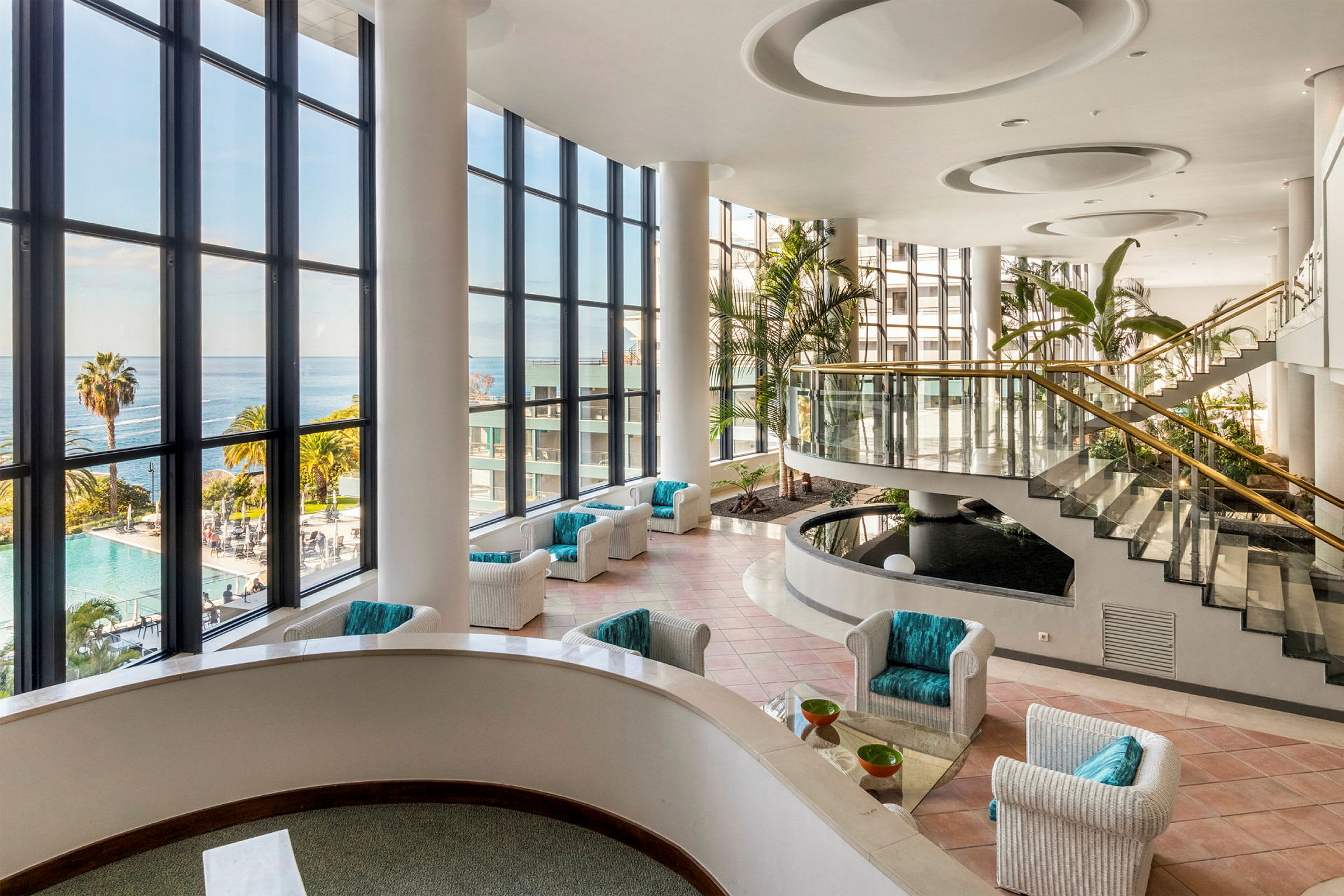 Hotellobby med stora glasfönster, utsikt över havet, sittplatser och trappor. (AI-genererad text)