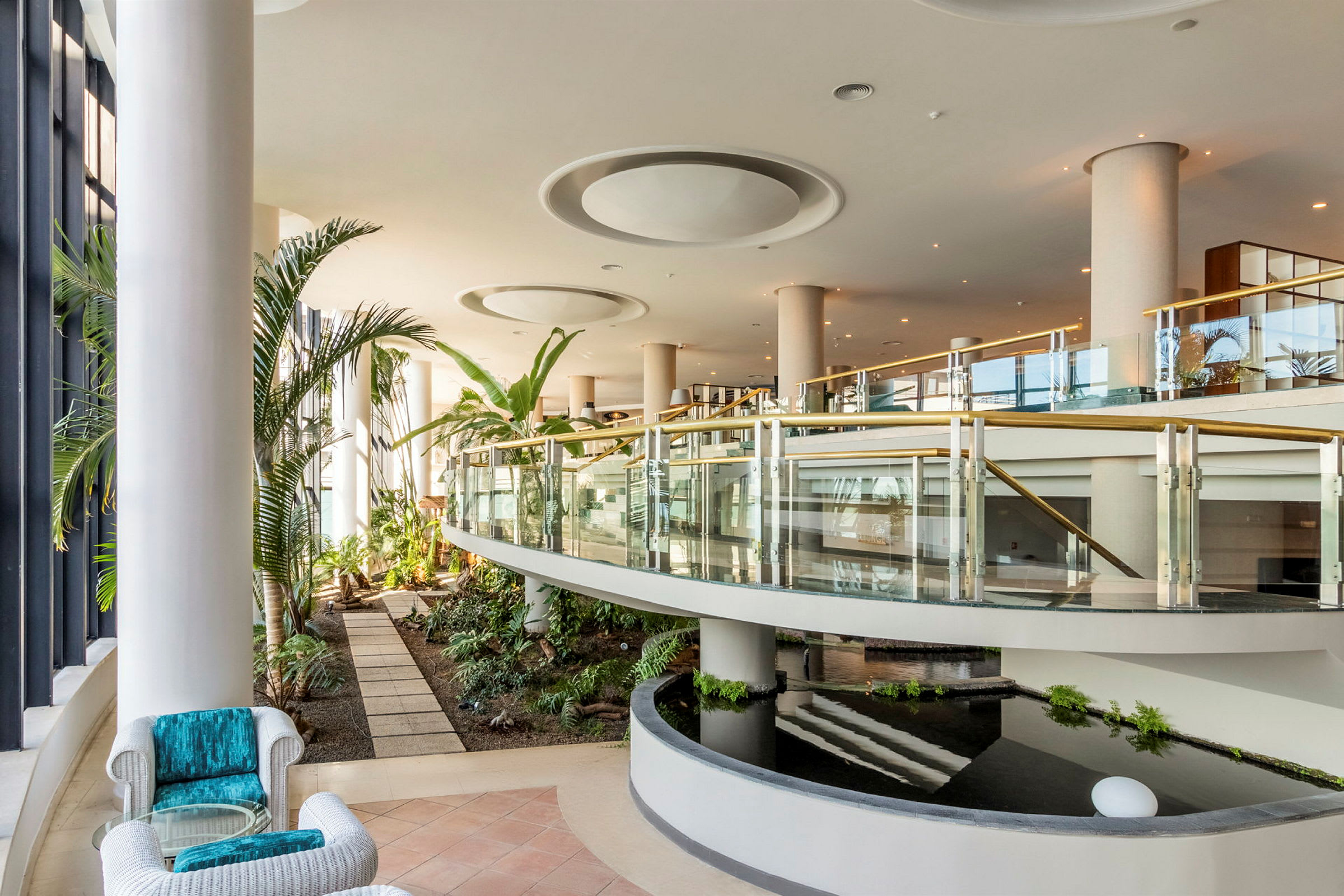En modern hotellobby med växter, glasräcken och vattenanläggning. (AI-genererad text)