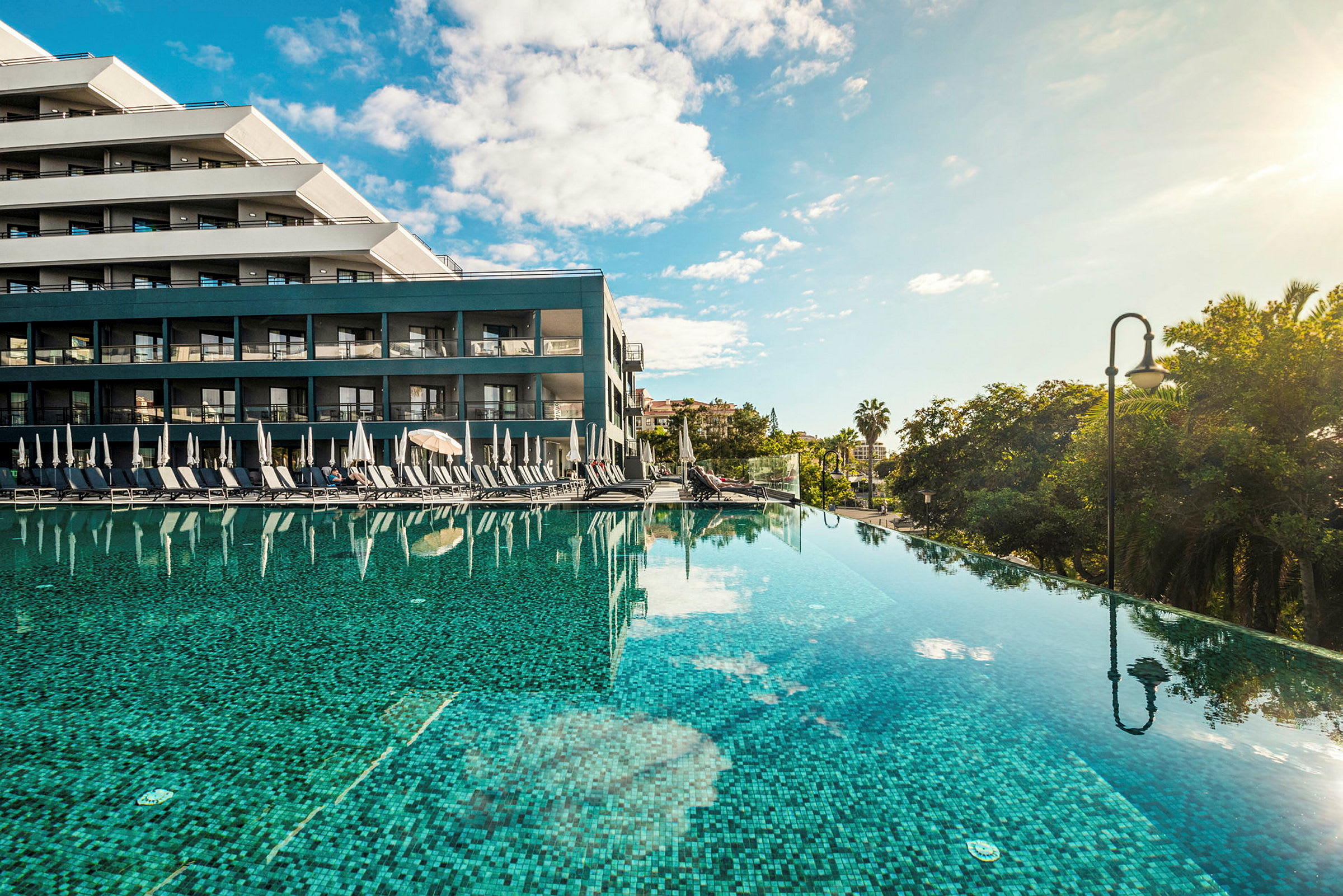 Ett hotell med pool, solstolar och parasoll vid kanten. Palmträd i bakgrunden. (AI-genererad text)