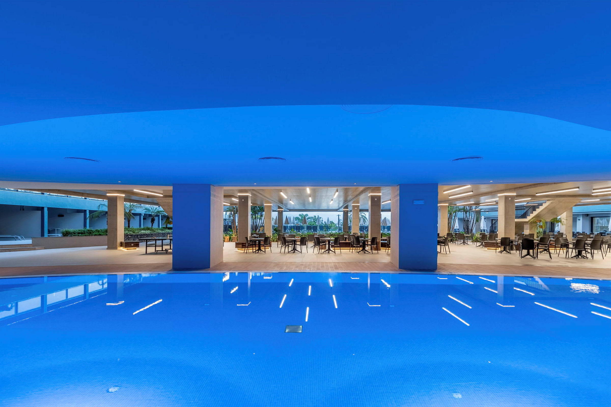 En modern pool med ljusblått vatten framför en öppen sittgrupp med bord och stolar. (AI-genererad text)