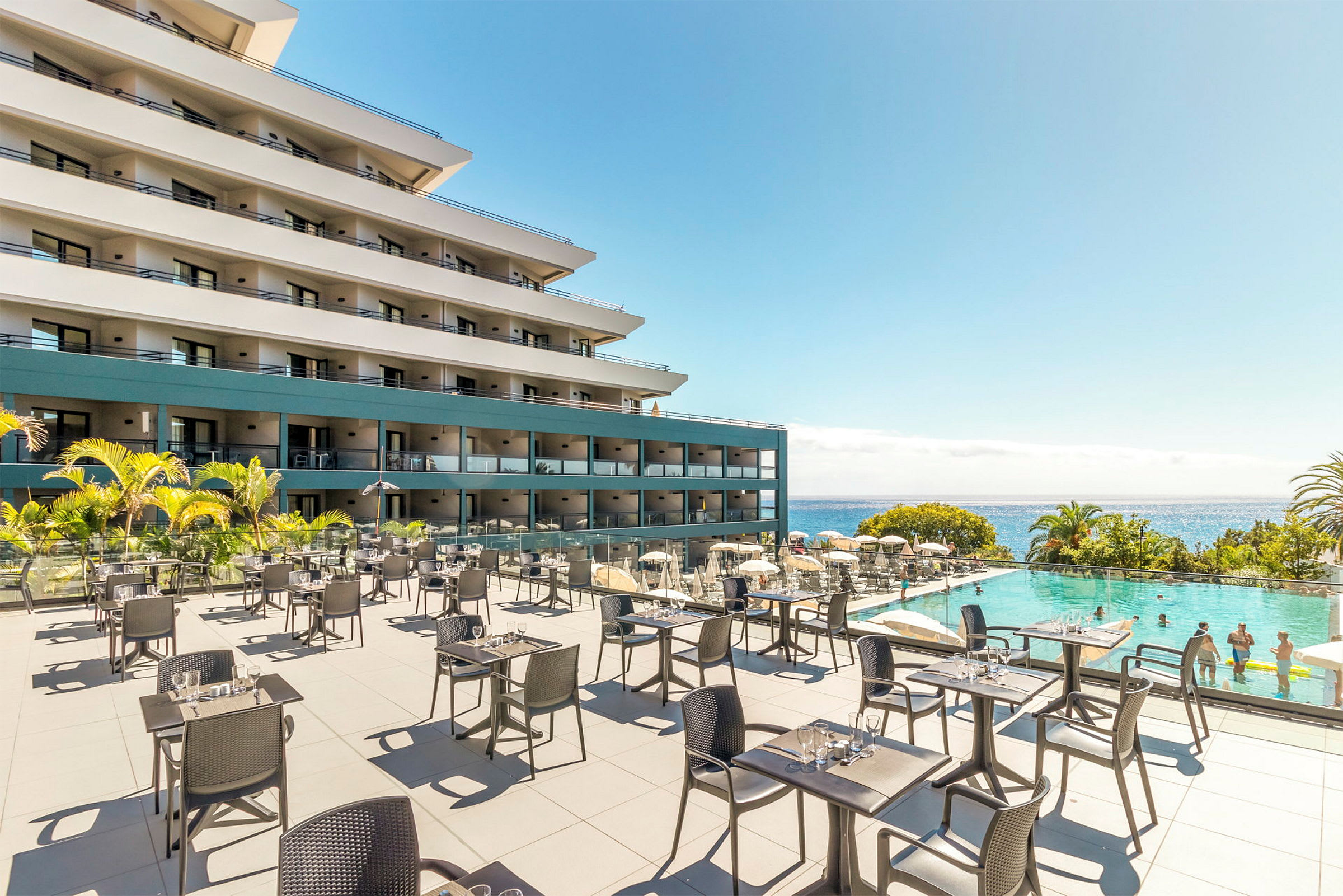 Uteservering med bord och stolar framför ett hotell och en pool med badgäster, med havet i bakgrunden. (AI-genererad text)