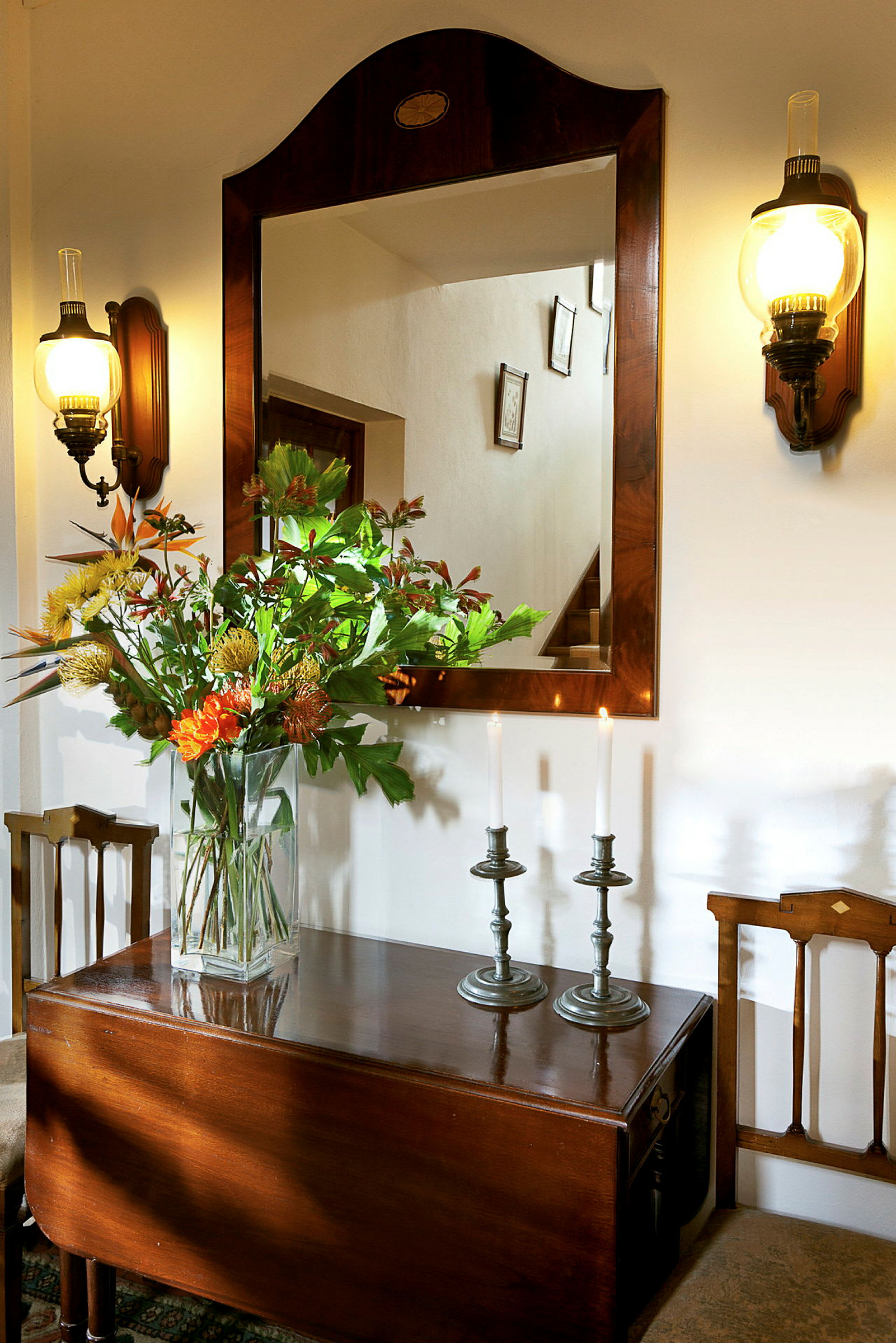 En spegel med träram, två vägglampor, och ett bord med en vas med blommor samt två ljusstakar med tända ljus. (AI-genererad text)